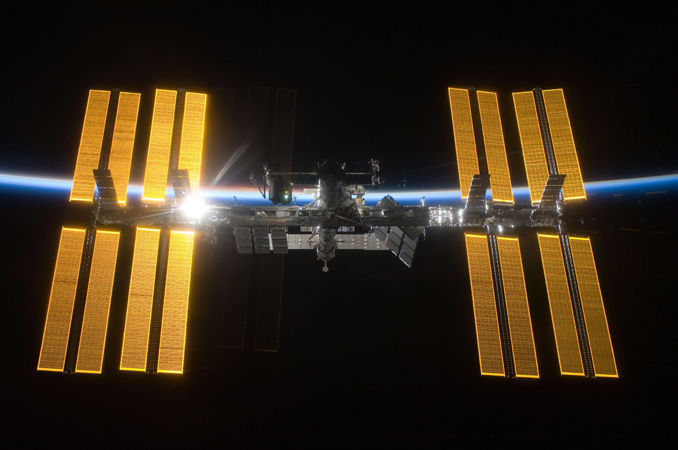 Auf diesem Bild sieht man die ISS vor der Nachtseite der Erde. Die aufgehende Sonne beleuchtet das dünne Band der Atmosphäre. 
Bild: NASA