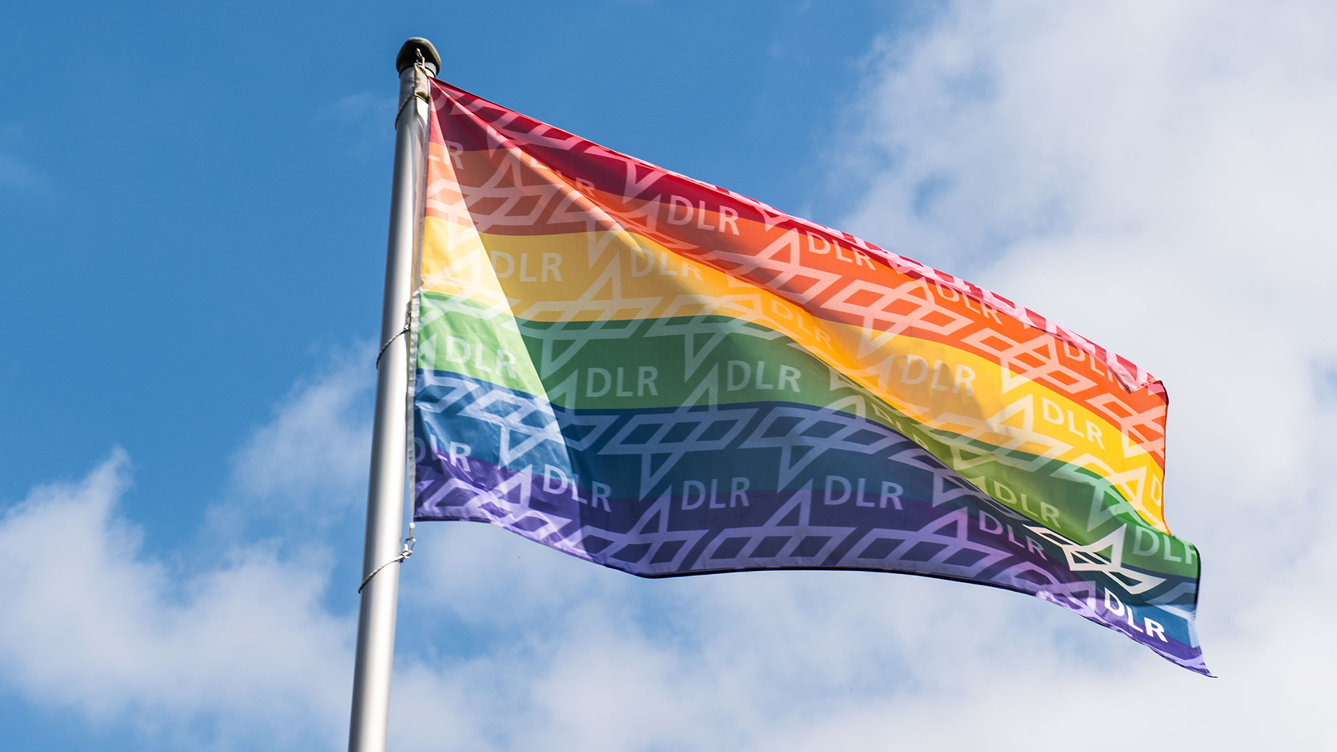 Gehisste Regenbogenflagge mit DLR-Logos vor blauem Himmel