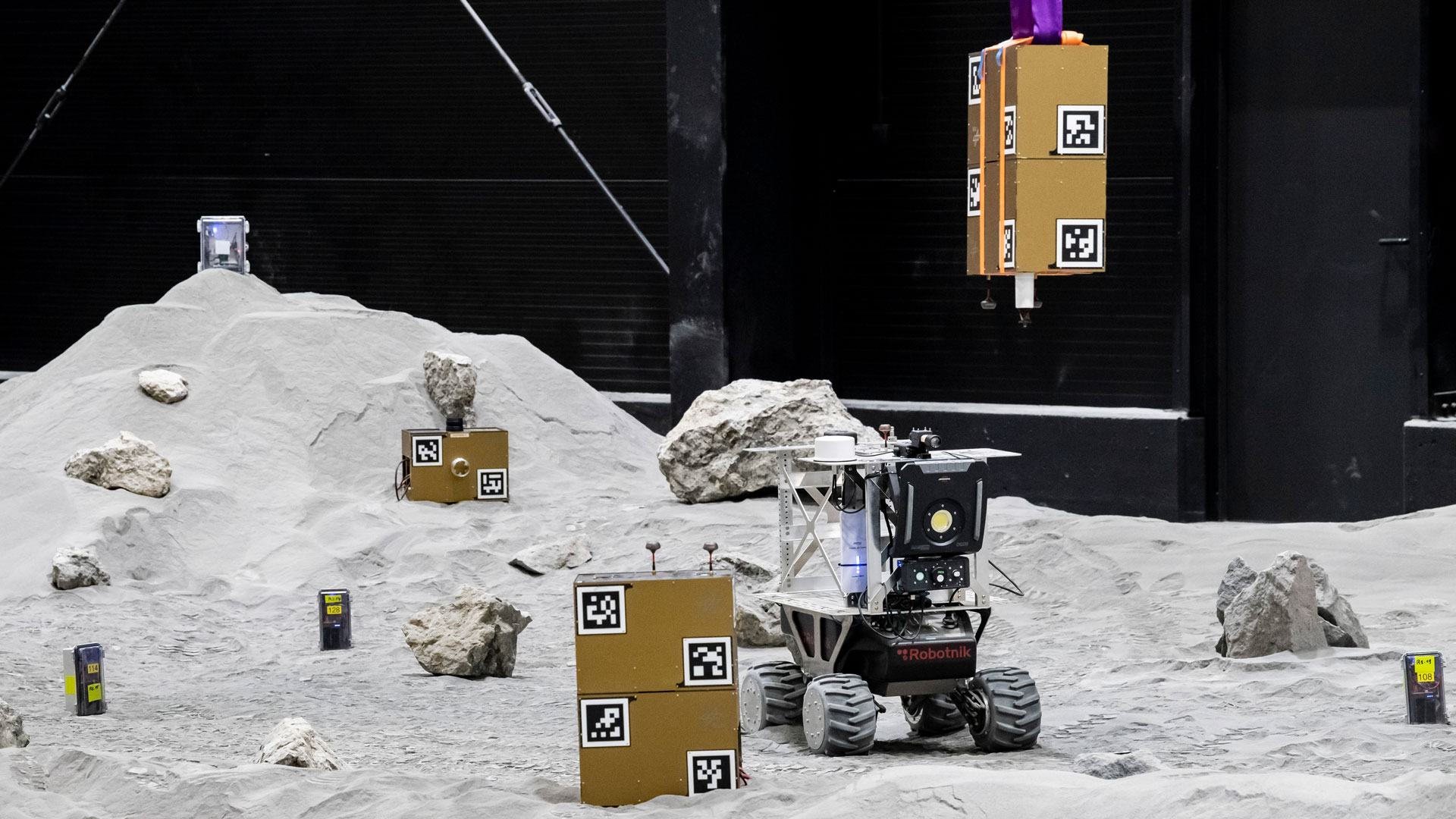 Das Bild zeigt goldfarbene Nutzlastboxen, weiße Sensoreier und einen Rover auf der grauen Regolith-Oberfläche der LUNA-Halle.
