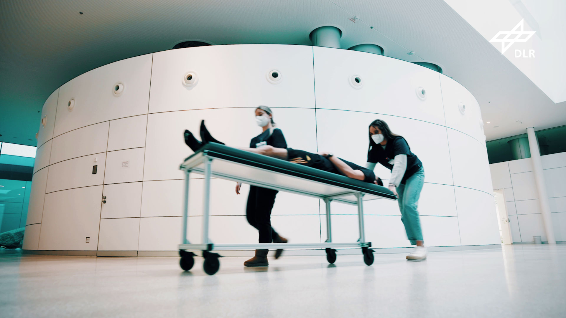 Den Weg von ihren Zimmern zu den Untersuchungen und Experimenten legen die Teilnehmenden in Kopftieflage auf einer Liege zurück. Ein Teilnehmer wird durch das Foyer des :envihab geschoben.