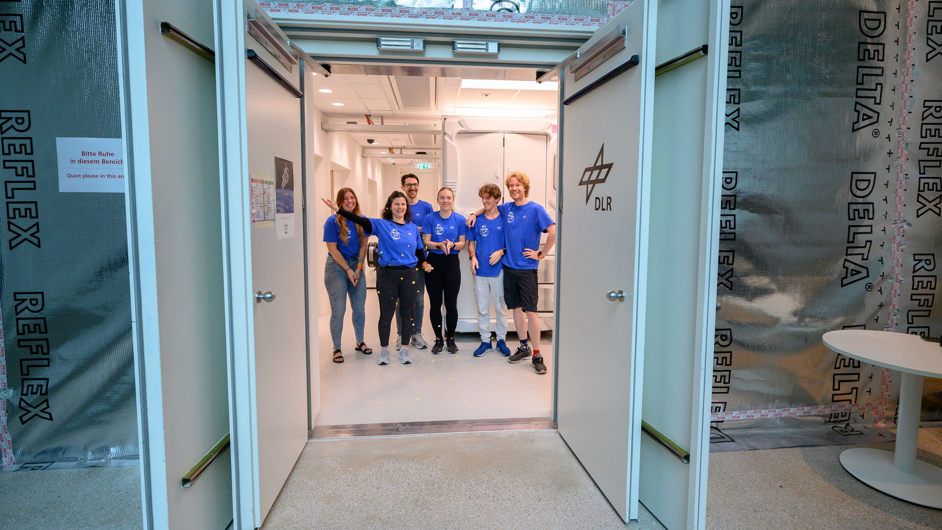 Drei Männer und drei Frauen sind durch geöffnete Flügeltüren zu sehen. Sie tragen blaue Mission-Shirts. Eine Frau wirft Konfetti.