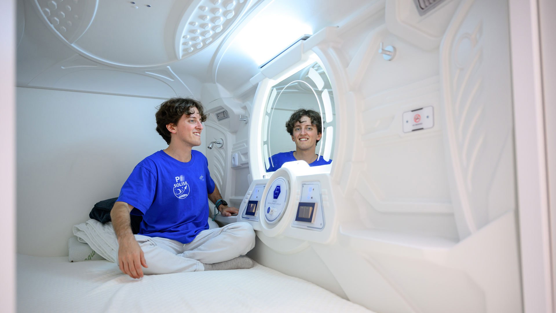 Ein junger Mann sitzt in seiner engen, weißen Schlafkapsel und schaut in einen Spiegel.