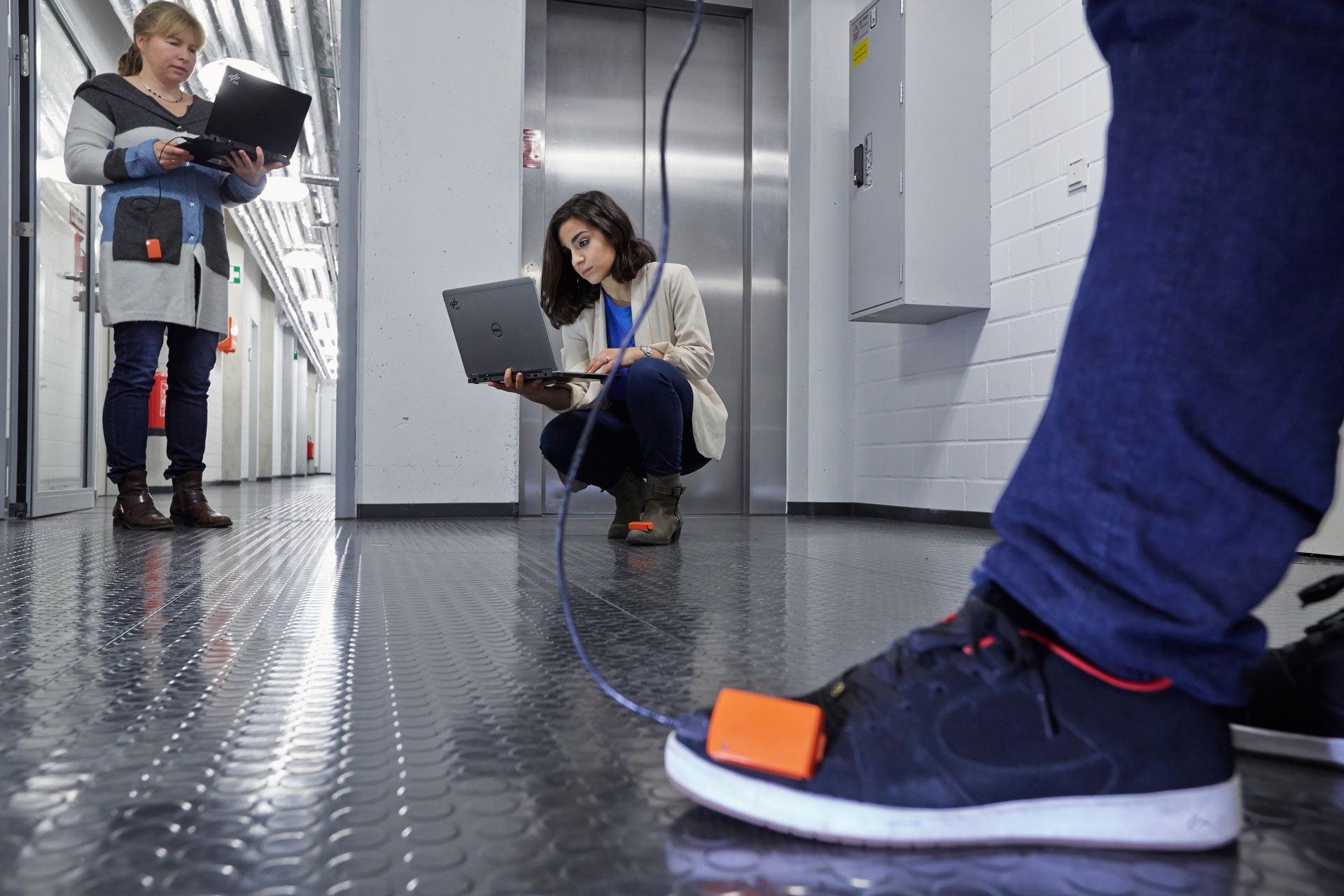 Das Bild zeigt ein kleines orangefarbenes Messgerät, das auf einem Schuh befestigt ist. Im Hintergrund arbeiten zwei Wissenschaftlerinnen an Laptops.