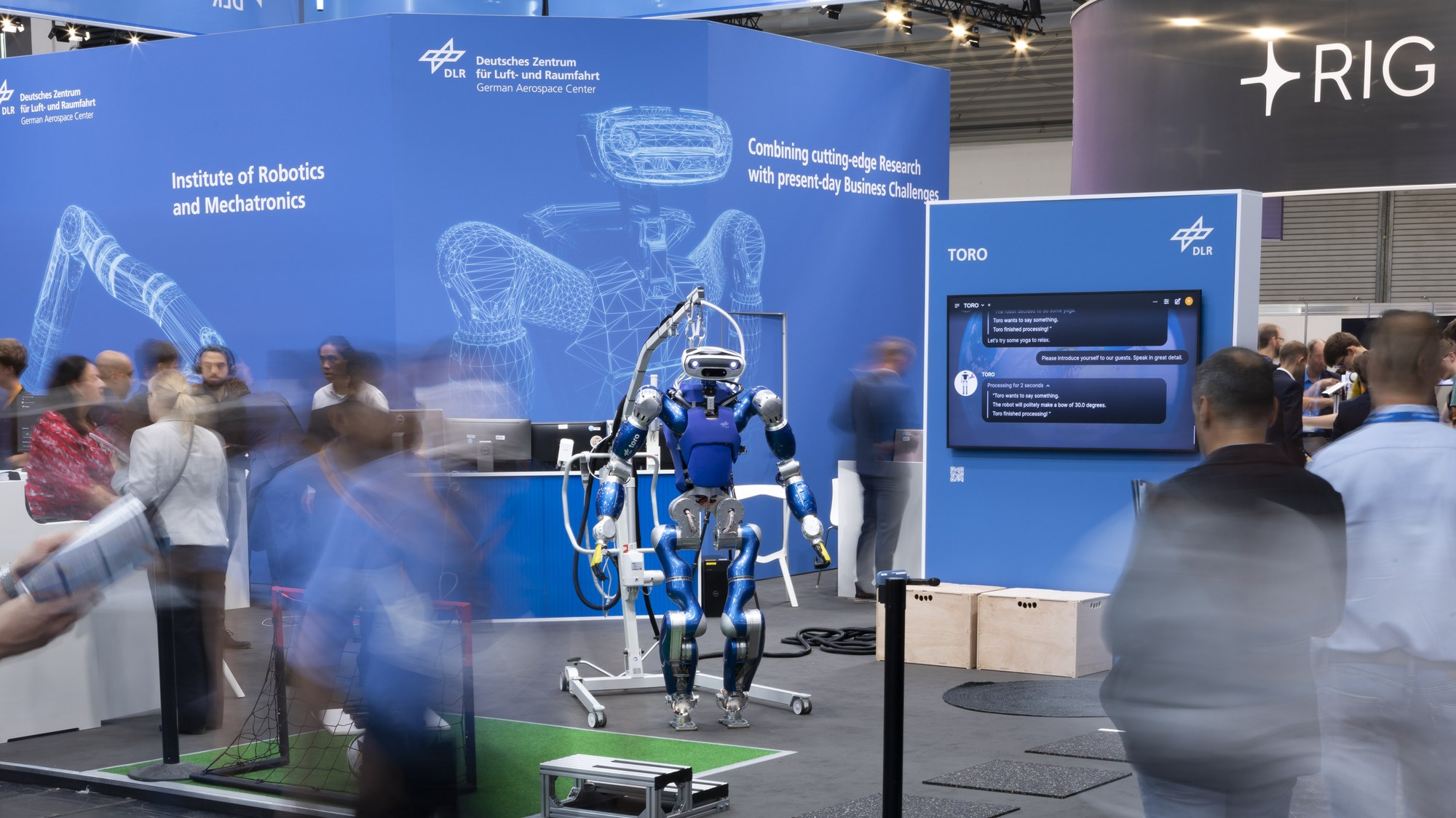 Das Bild zeigt den zweibeinigen DLR-Roboter TORO. Der Roboter ist blau und silberfarben und hebt einen Arm.
