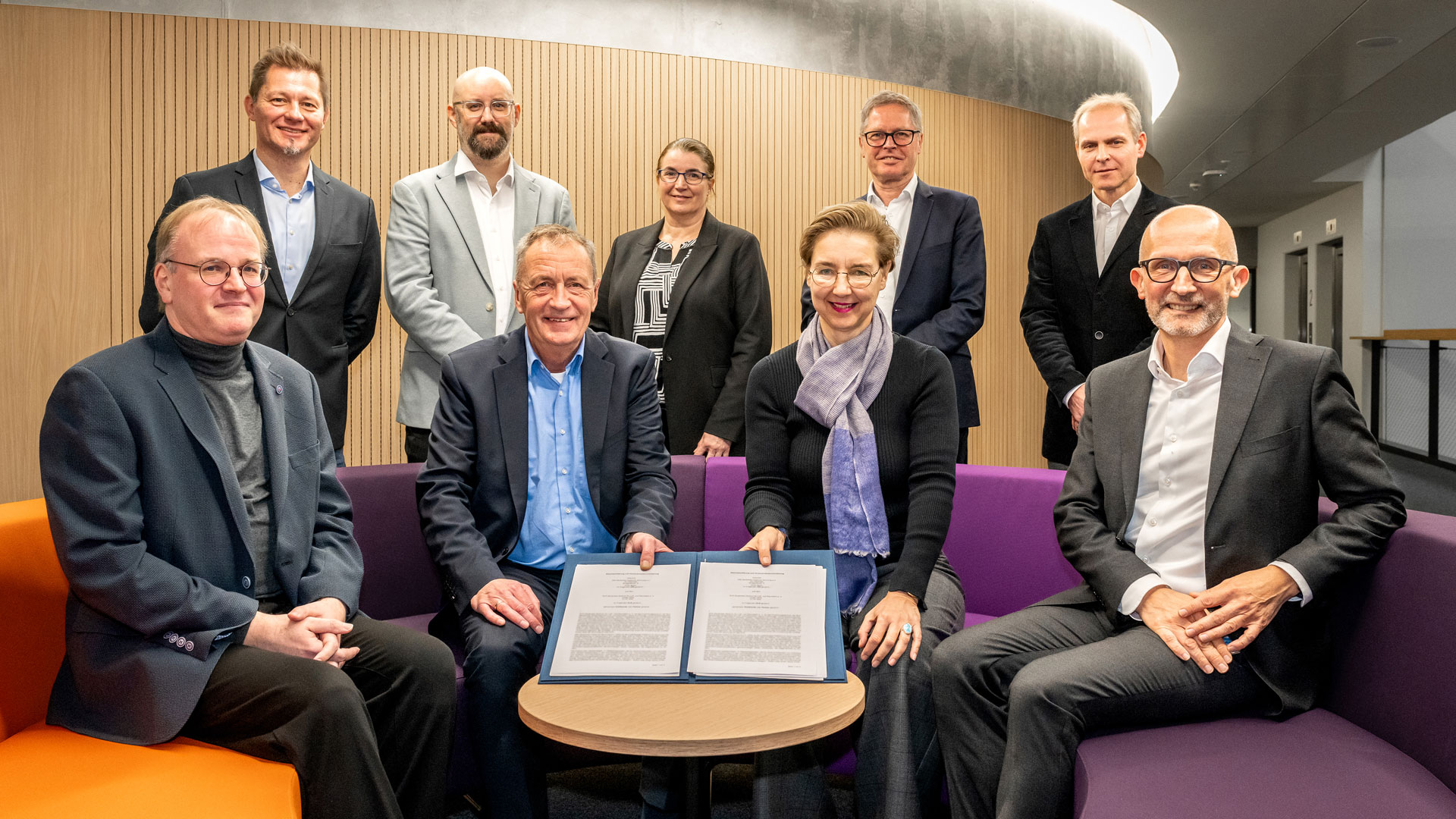 Das Bild zeigt neun Personen von DLR, DIN und DLR Projektträger. Die Verantwortlichen von DLR und DIN in der Mitte halten die unterzeichnete Vereinbarung.