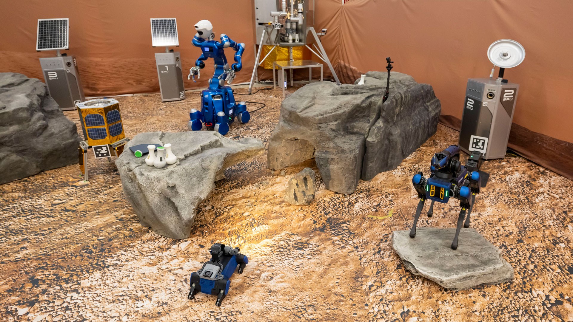 Das Bild zeigt drei unterschiedliche Roboter in einer rötlichen Umgebung, die der Mars-Oberfläche nachempfunden ist.