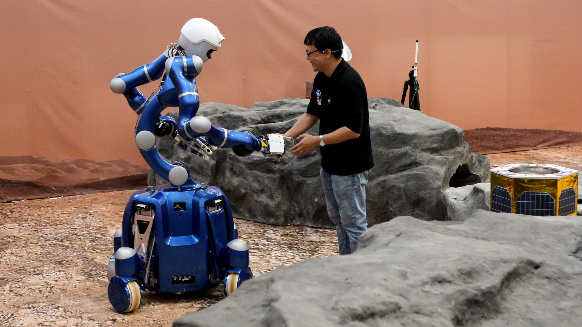 Ein blauer, humanoider Roboter und ein Wissenschaftler reichen sich die Hand. Sie stehen in einer rötlichen Umgebung, die der Mars-Oberfläche nachempfunden ist.