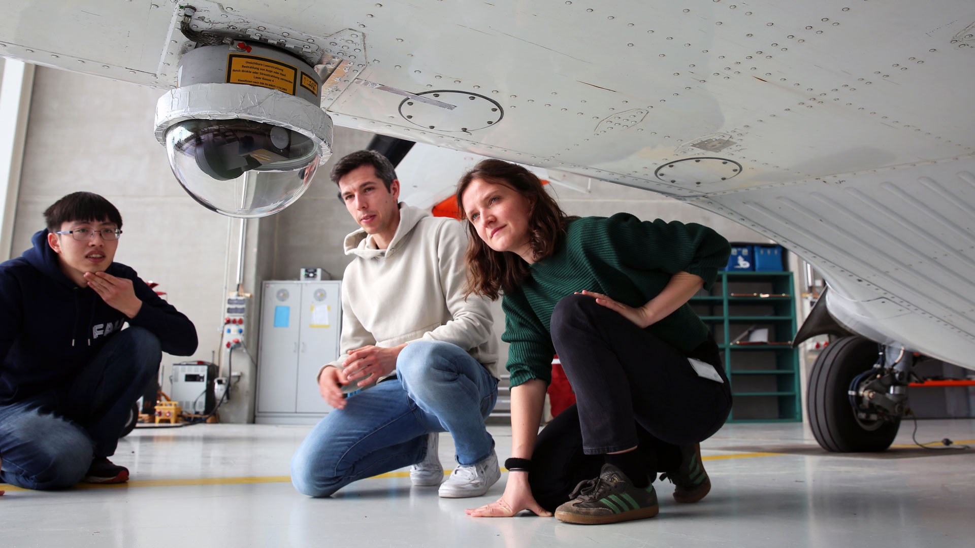 Drei junge Forschende hocken unter dem Rumpf eines kleinen weißen Flugzeugs. Sie blicken auf ein technisches Element, das am Rumpf angebracht ist.