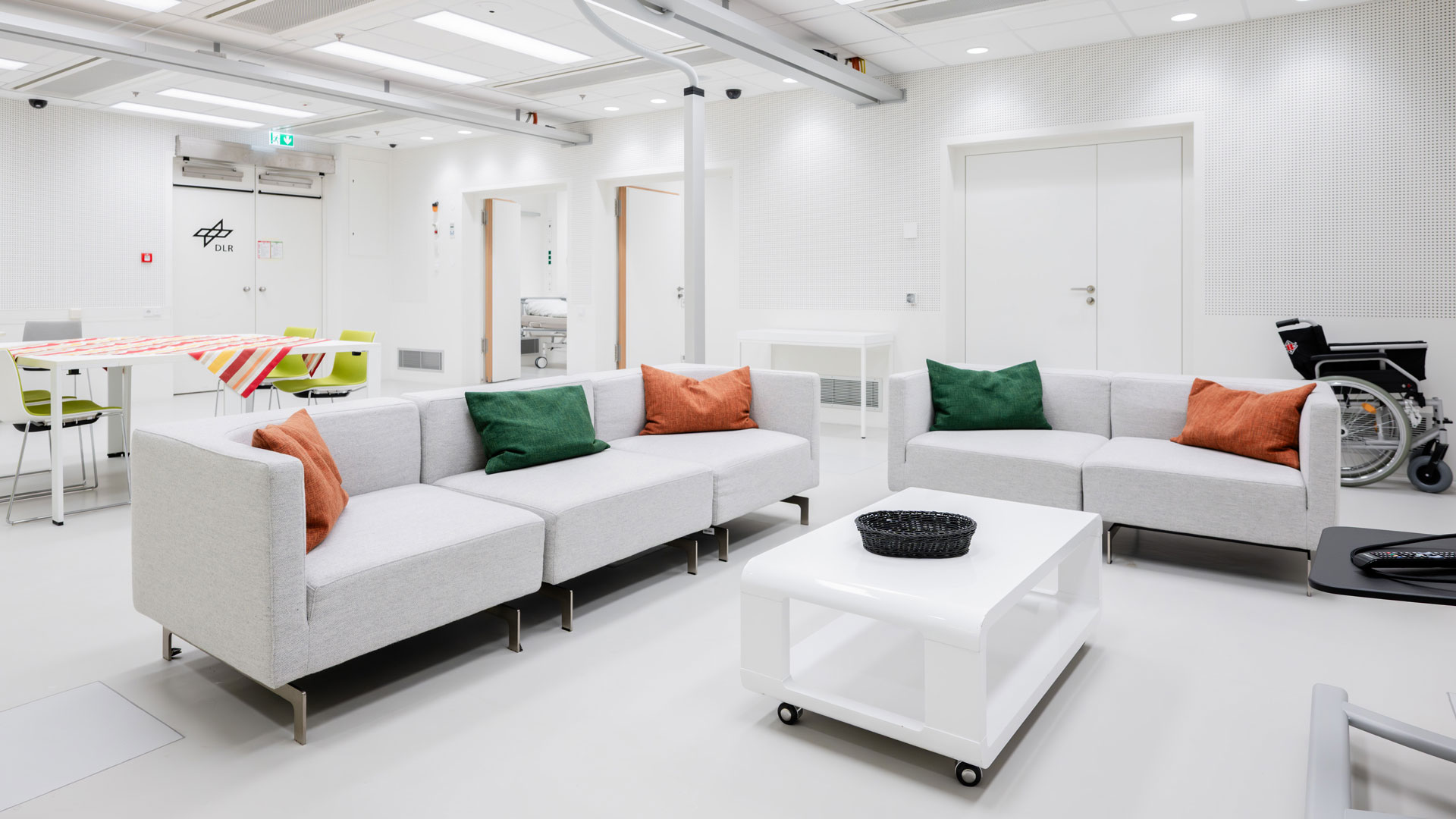 Auf dem Bild ist ein weißes Sofa mit bunten Kissen zu sehen, im Hintergrund ein Tisch und Nebenräume. Es handelt sich um den Wohnbereich M5 im :envihab.