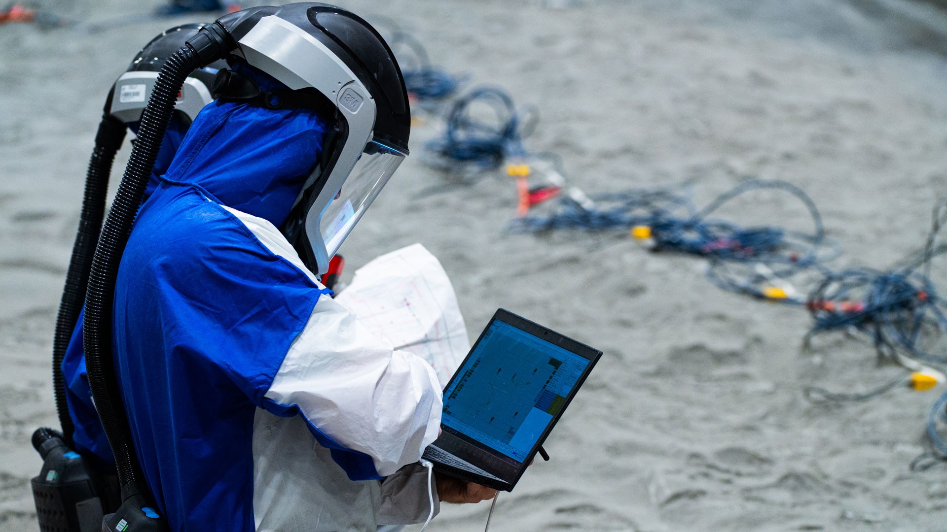 Eine Person im weiß-blauen Schutzanzug mit Atemschutz hält ein Tablet. Im Hintergrund sind Kabel zu erkennen.