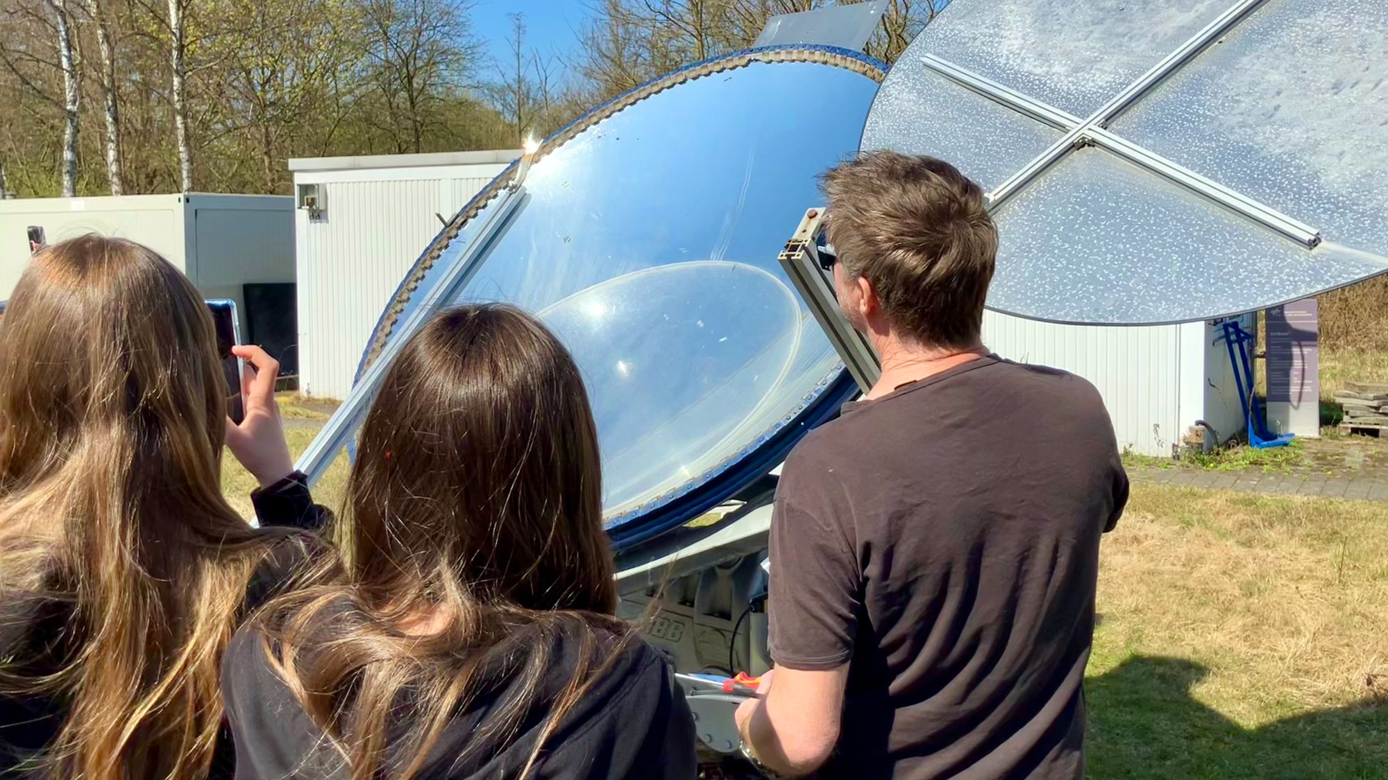 Schülerinnen und ein Wissenschaftler stehen vor einem Diskus-Spiegel. Davor wird ein Punkt sehr hell.