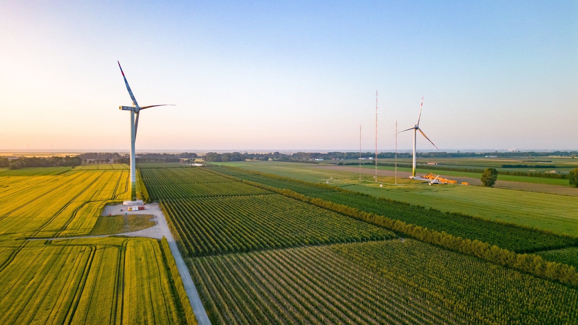 Zwei Windräder mit viel grüner Fläche. Im Hintergrund Sonnaufgang oder Sonnuntergang.