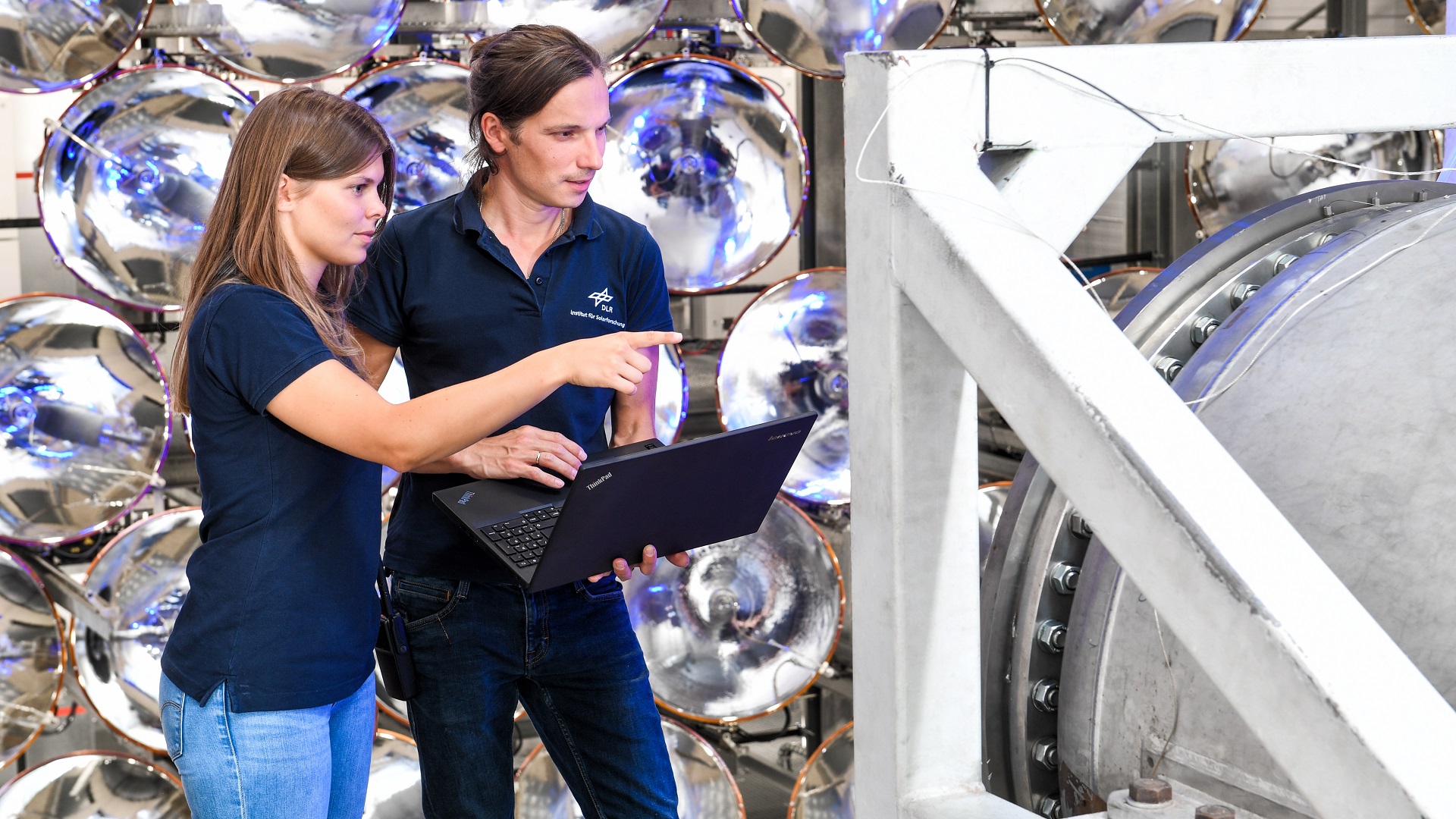 In einem Labor der Solarforschung stehen zwei DLR Mitarbeitende. Links, eine Frau in dunkelblauen Poloshirt und rechts von ihr ein Mann in dunkelblauen Shirt. Er hält einen Laptop in der Hand und sie zeigt auf das turbinenartige Gerät.