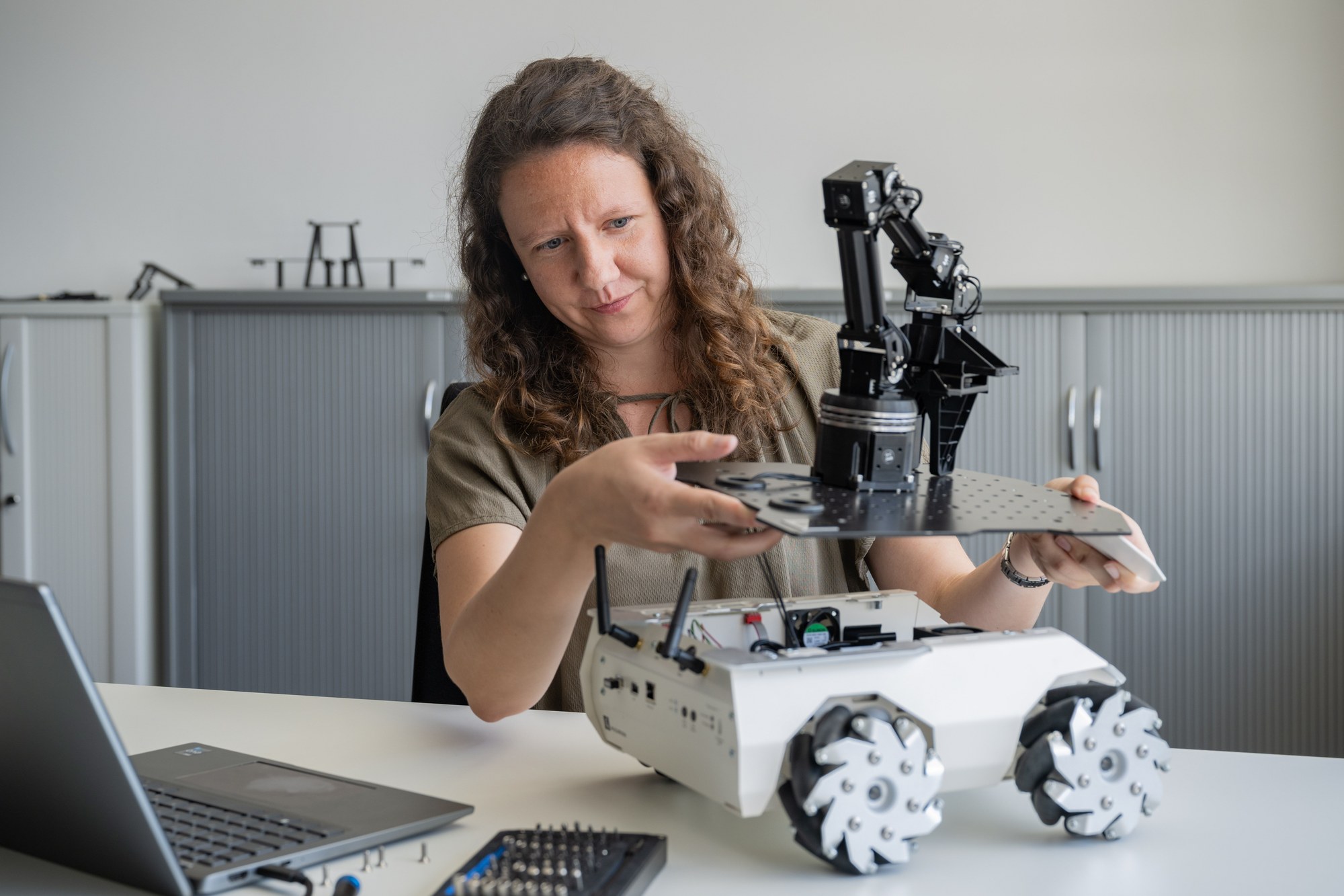Junge Frau sitzt an ihrem Arbeitsplatz und schraubt an einem Roboter, der auf dem Tisch steht.