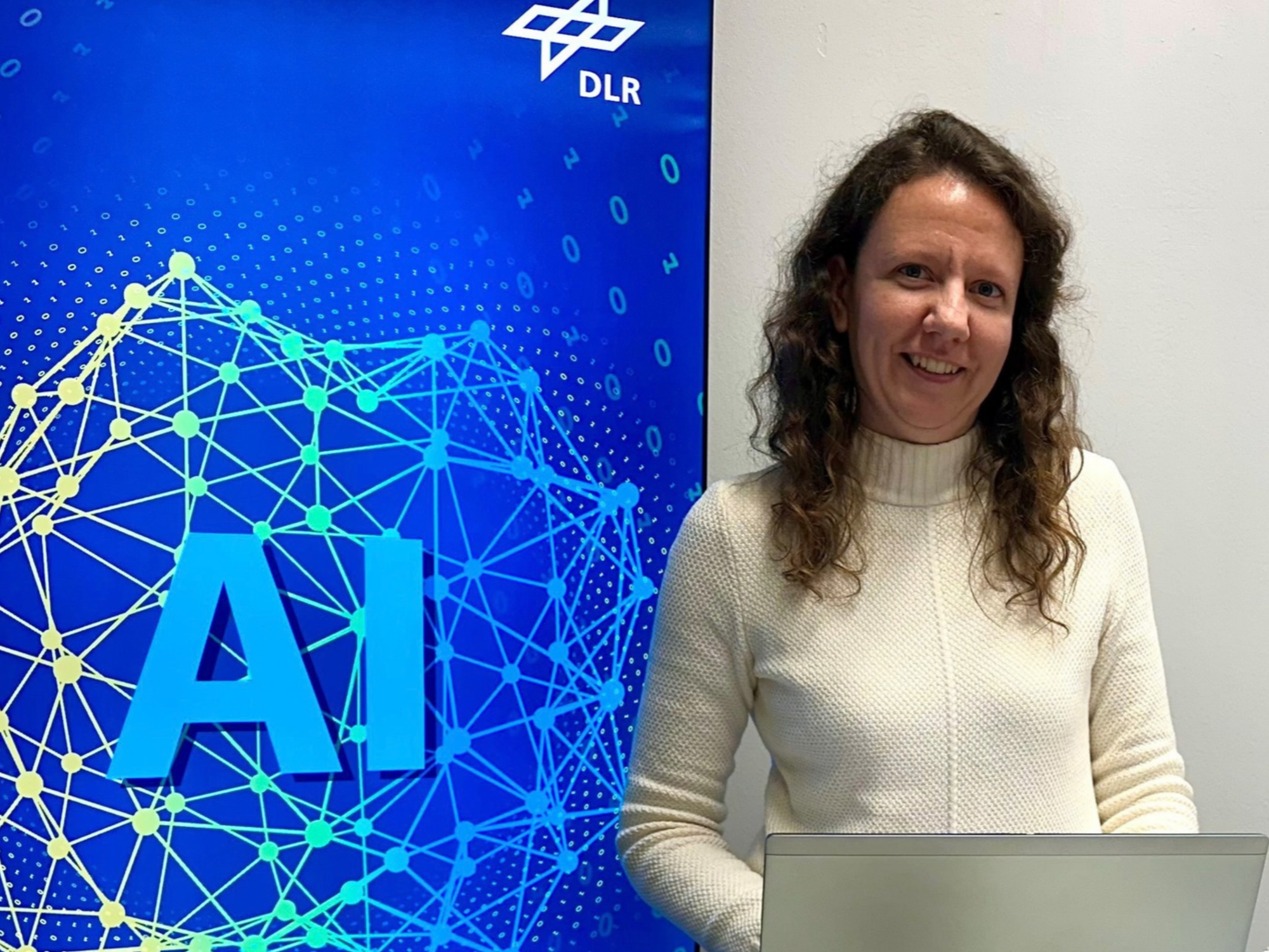 Junge Frau steht mit ihrem Laptop vor einer Wand auf der AI steht und ein neuronales Netz abgebildet ist.