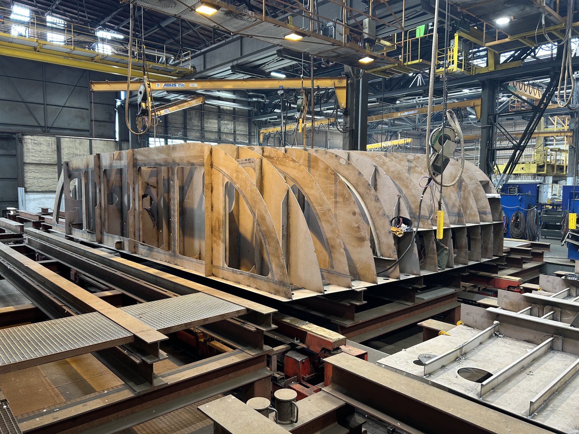 Eine Werfthalle mit Kränen, Kabeln und Gerüsten an den Seiten und der Decke, in der Mitte ist eine Sektion aus Stahlplatten, die nebeneinander in gleicher Größe angeordnet sind und an der oberen Seite abgerundete Ecken hat