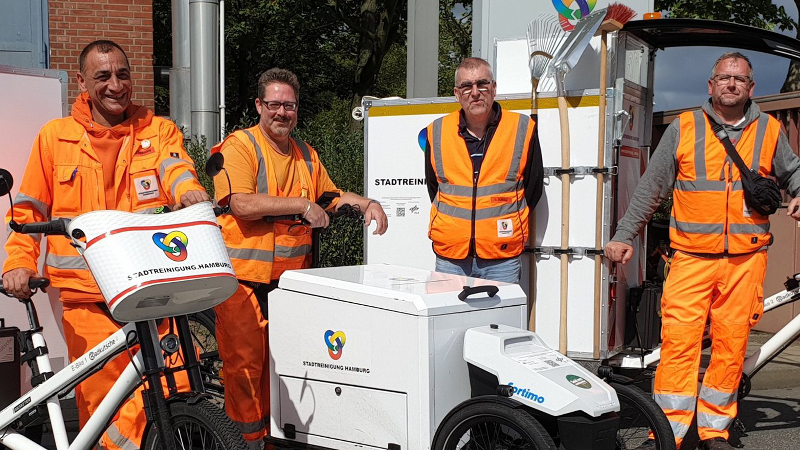 Zu sehen ist eine Gruppe von vier Mitarbeitern der Stadtreinigung Hamburg in Arbeitskleidung. Sie stehen um drei verschiedene Modelle von Lastenrädern herum. Die Lastenräder sind auf die Nutzungsanforderungen der Stadtreinigung ausgelegt.