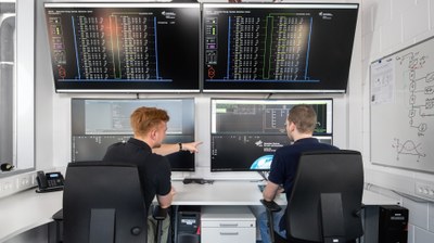 Zwei Personen sitzen an einer Leitwarte vor mehreren Großbildschirmen und weisen auf einen der Monitore.