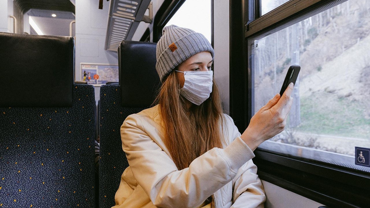 Eine Frau sitzt mit Maske in einem Zug und hält ihr Handy in der Hand.