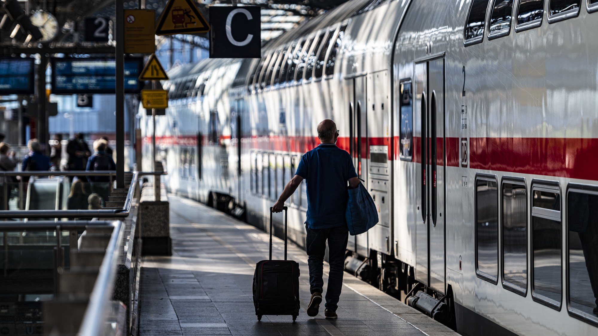 Ein Passagier am Kölner Hauptbahnhof geht mit Rollkoffer den Bahnsteig entlang. Am rechten Arm trägt er eine blaue Tasche. Am Gleis steht ein Intercity mit geschlossen Türen zur Abfahrt bereit.