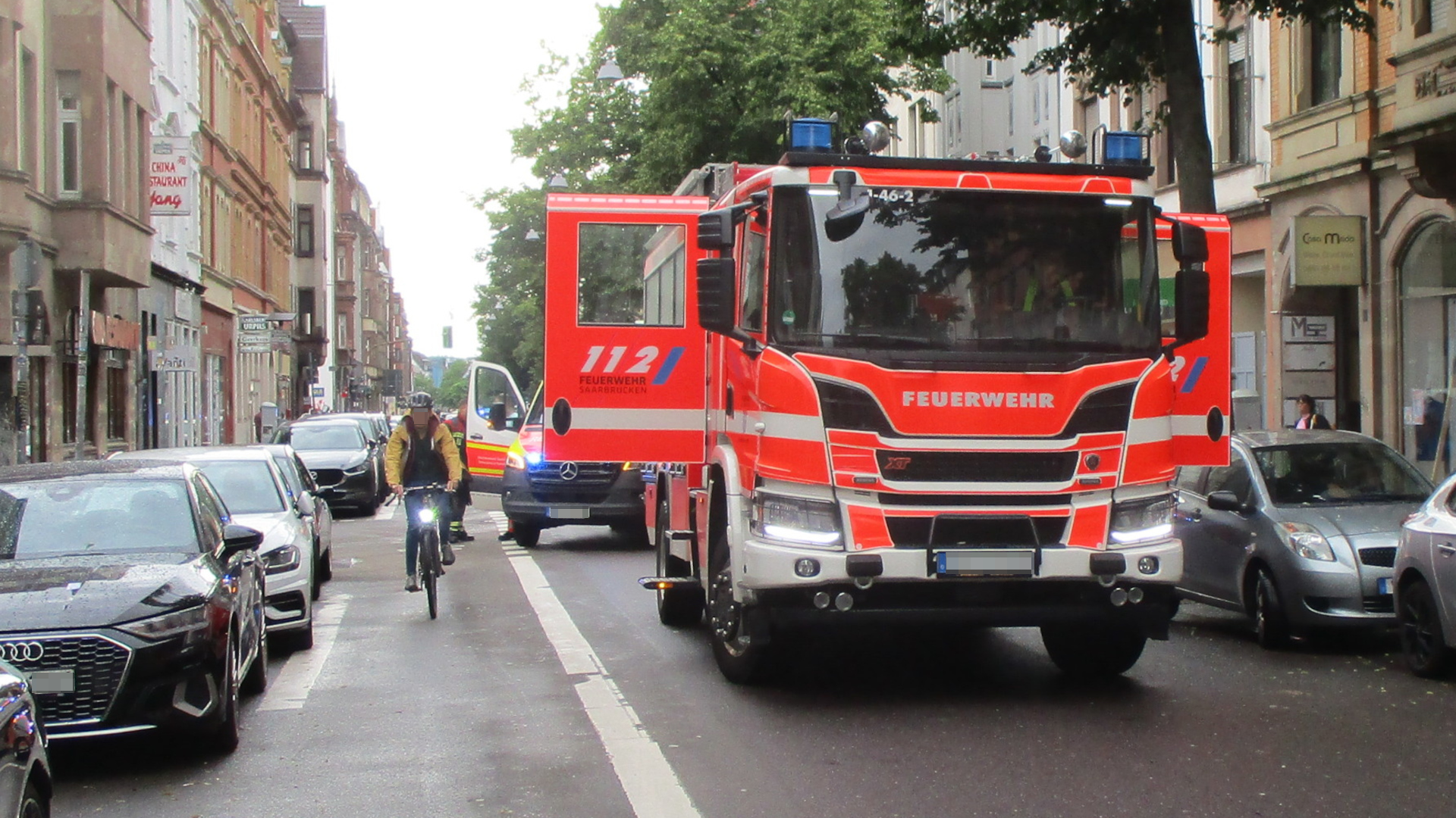 Einsatzfahrzeuge der Feuerwehr stehen auf einer Einbahnstraße. Ein großes Gerätefahrzeug sowie ein kleinerer Bus sind hintereinander auf der Fahrbahn abgestellt. Auf der Straße ist links ein Fahrradweg abgeteil, auf dem ein Radfahrer der Kamera entgegenfährt. Auf beiden Straßenseiten stehen Pkw in Parkplätzen. Rechts reihen sich Bäume am Gehweg entlang. Die Straße ist von Altbauten gesäumt..