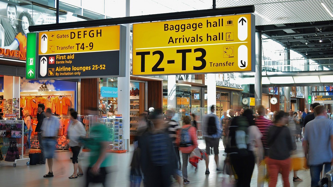 Eine Zeitrafferaufnahme von Personen an einem Terminal im Flughafen. Zu erkennnen ist die große Beschilderung und kleine Läden im Hintergrund.