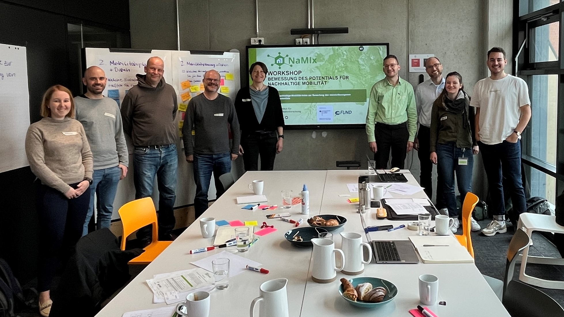 Abgebildet ist eine Gruppe von neun Personen unterschiedlichen Alters und Geschlechts. Sie stehen freundlich in die Kamera lächelnd hinter einem Tisch mit Zetteln, Stiften, Getränken und Backwaren. Hinter ihnen sind Pinnwände und ein Monitor erkennbar, auf dem eine Präsentation zum Projekt dargestellt wird.