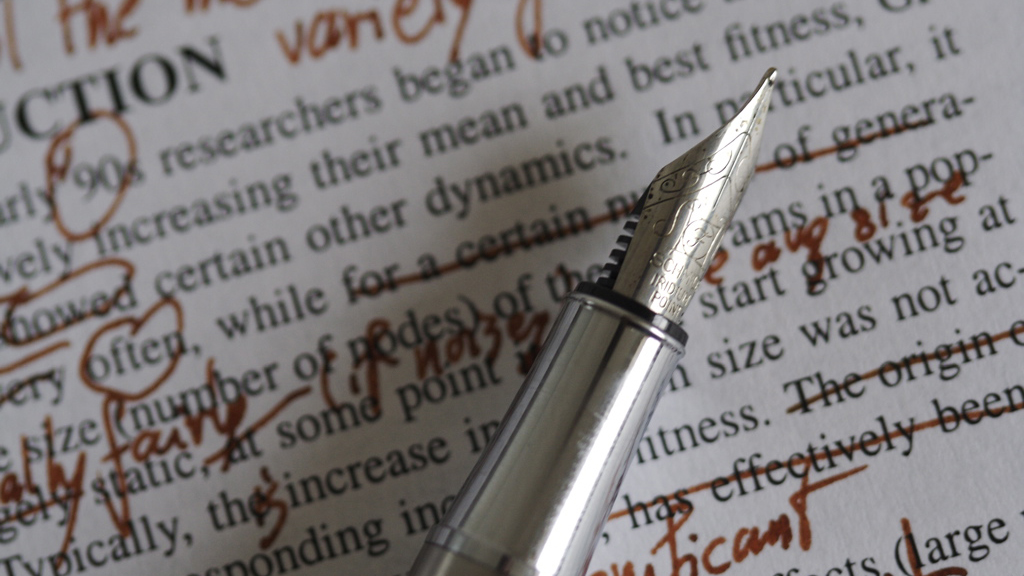 Zu sehen ist ein Füller mit roter Tinte, auf einem bedruckten Papier auf dem handschriftlich Korrekturen vorgenommen wurden.