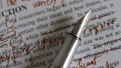 Zu sehen ist ein Füller mit roter Tinte, auf einem bedruckten Papier auf dem handschriftlich Korrekturen vorgenommen wurden.