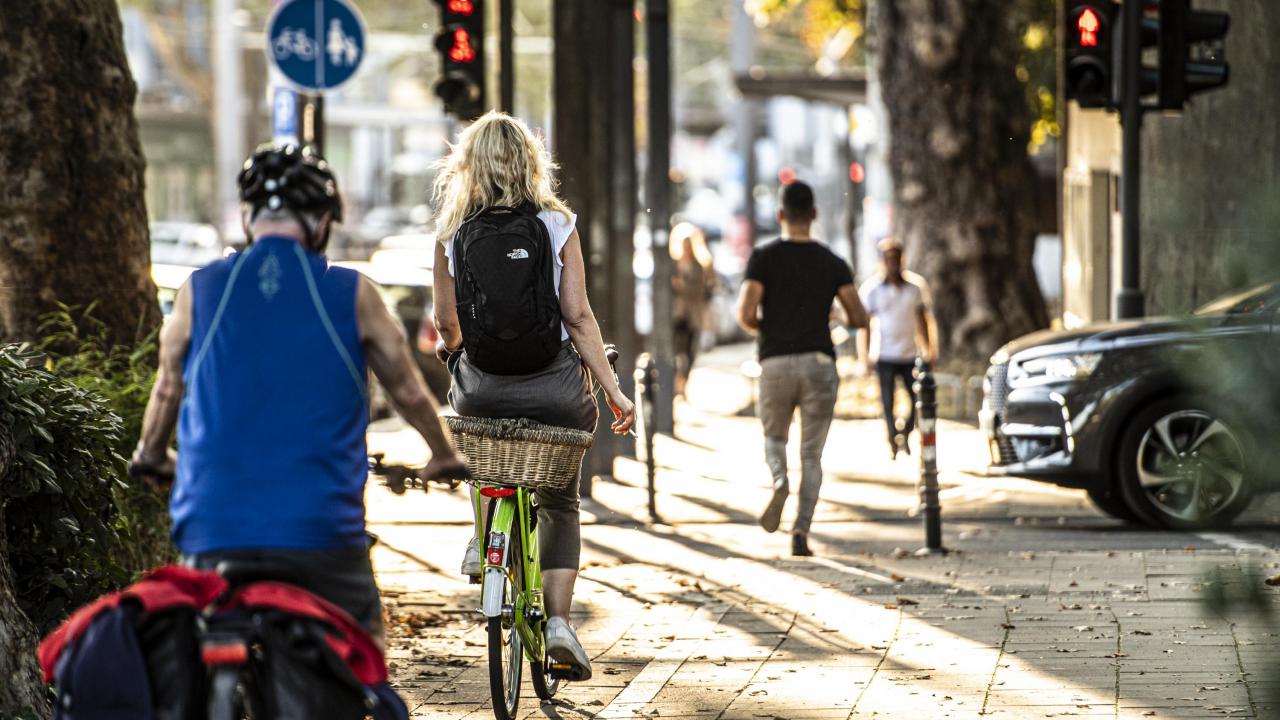Zu sehen ist ein Fahrradweg in einer Stadt. Mehrere Personen fahren über diesen Weg. Ein Auto möchte den Weg kreuzen.