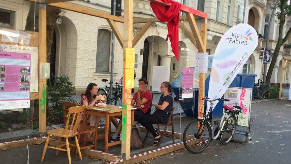 Ein Pavillion-Holzkonstrukt auf der Straße, indem Menschen sitzen.