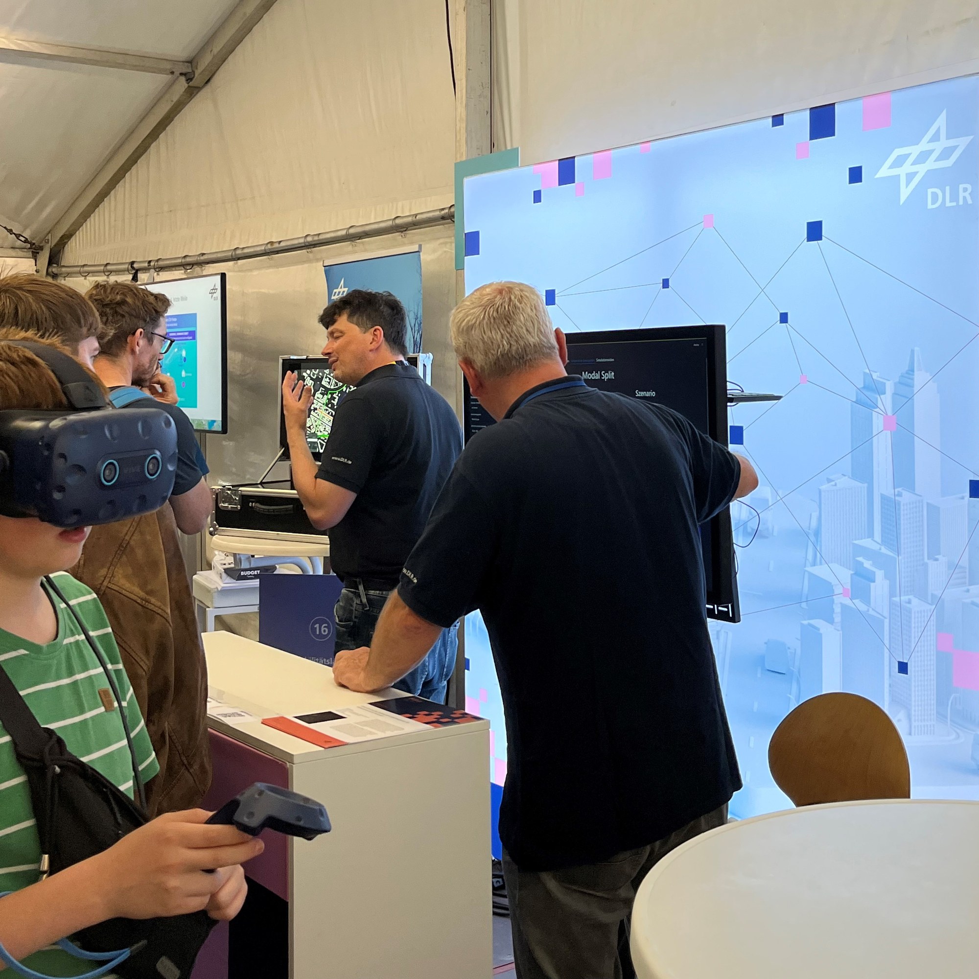 Zwei Wissenschaftler präsentieren vor Bildschirmen verschiedene Simulationen. Ein Kind hat eine VR-Brille auf.