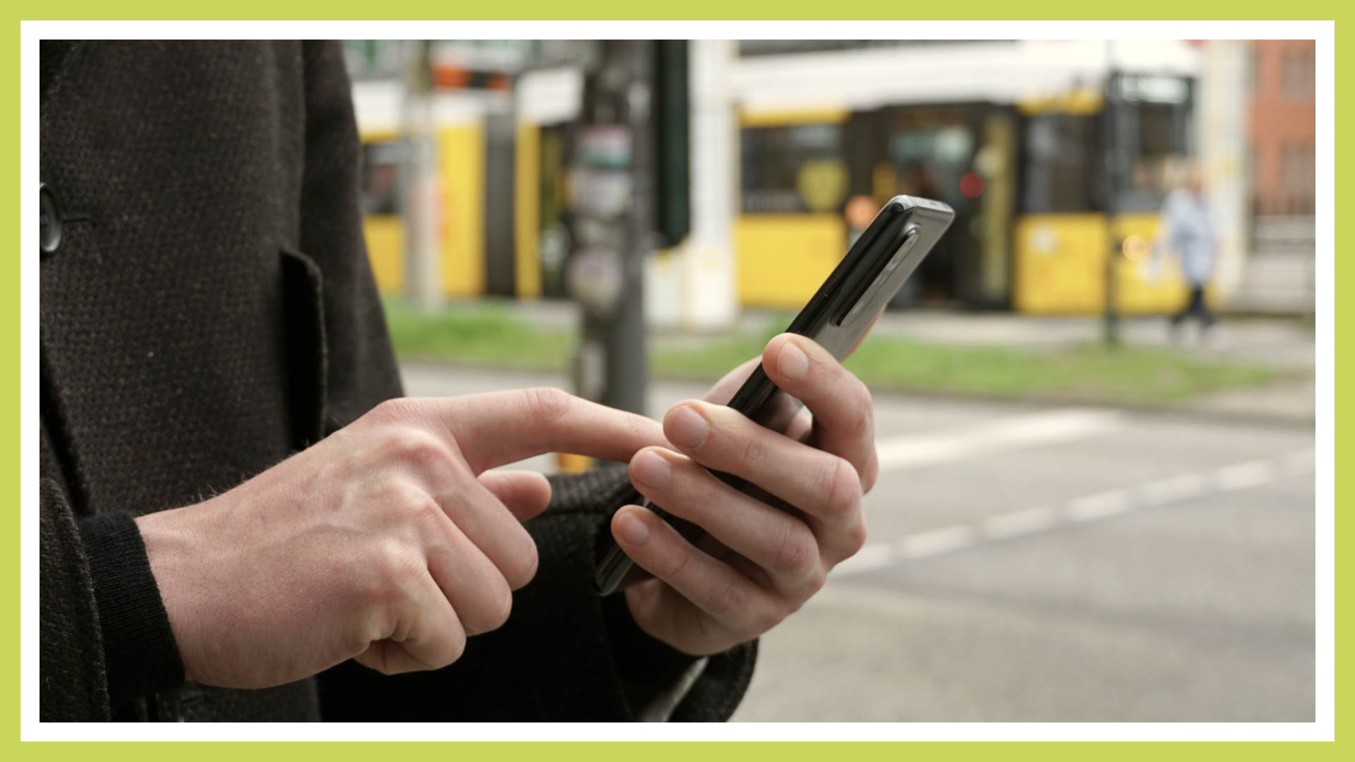 Zu sehen ist ein Foto einer Person die ein Smartphone bedient. Im Hintergrund fährt eine Straßenbahn.