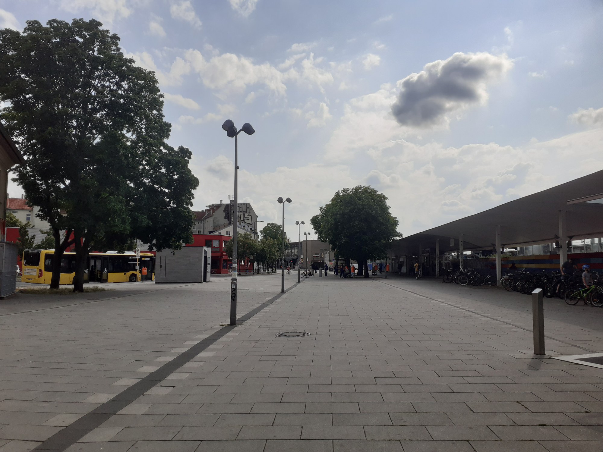 Ein großer, grau bepflasterter Platz ist im Vordergrund. In der Ferne sind entfernt Busstationen erkennbar.