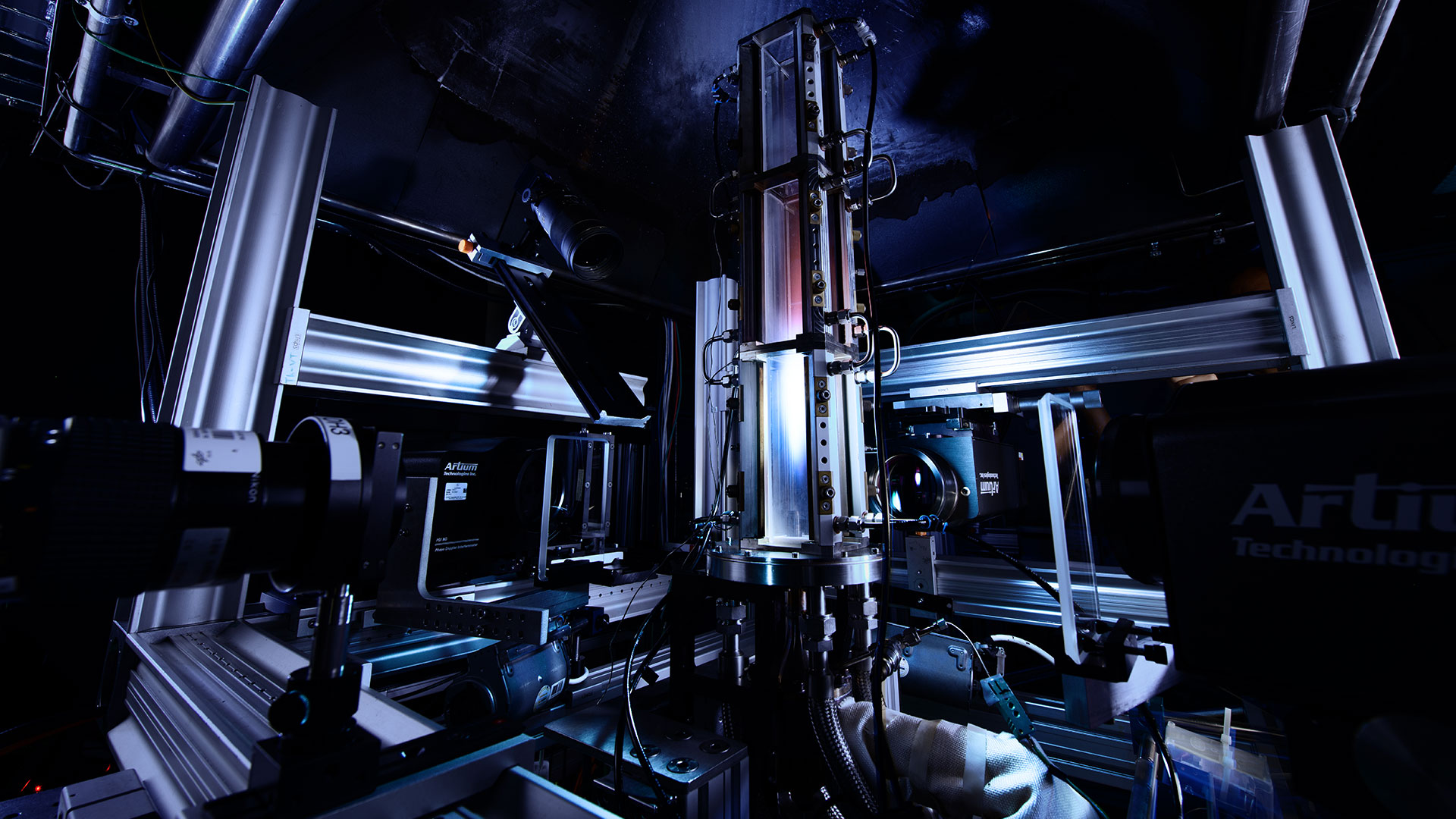 Das Bild zeigt in der Mitte eine große, optisch zugängliche Brennkammer eines atmosphärischen Verbrennungsprüfstands. Rings um die Brennkammer sind mehrere Kamera- und Lasersysteme aufgebaut, die für Messungen genutzt werden können.