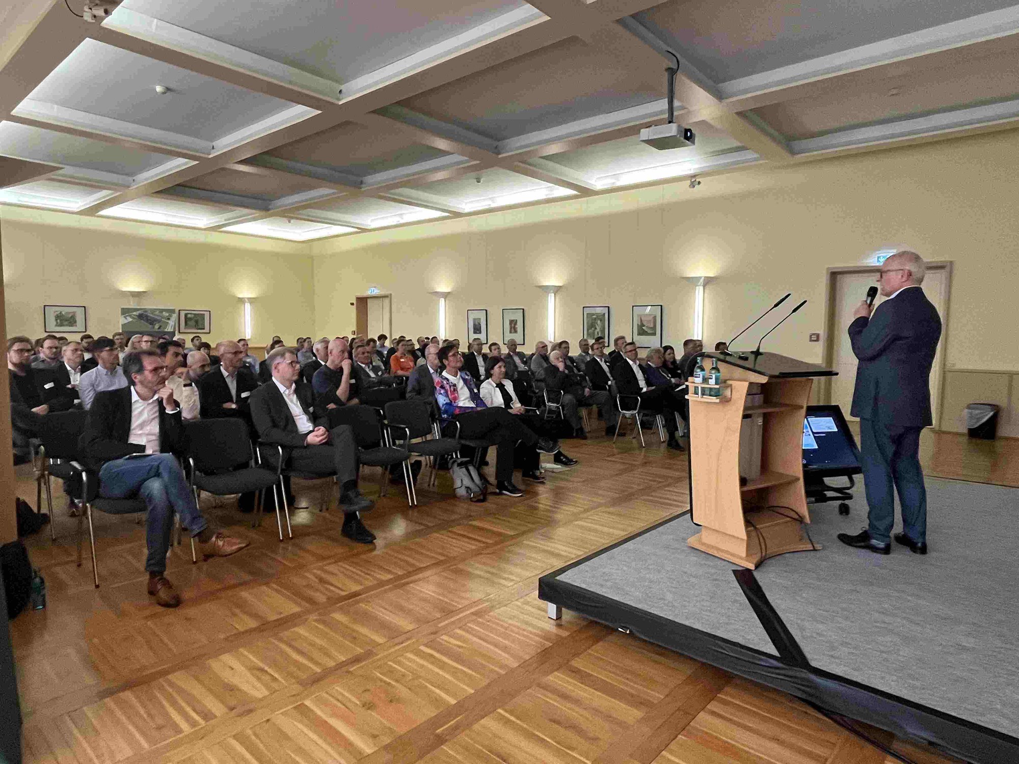 Peter Wehle steht mit Mikrofon auf der Bühne rechts und spricht zu einer Gruppe von rund 100 Menschen, die in einem festlichen Saal vor ihm auf Stuhlreihen sitzen.