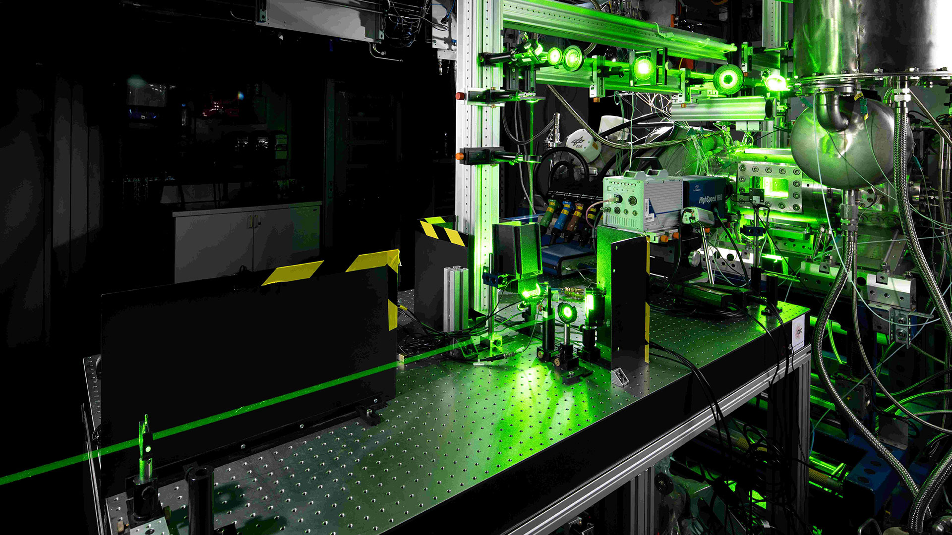 Das Bild zeigt den Hochdrückbrennkammerprüfstand HiPOT. Hinten rechts ist die optisch zugängliche Brennkammer zu sehen, in der Bildmitte ein komplexes optisches System für die eingesetzte Lasermesstechnik.