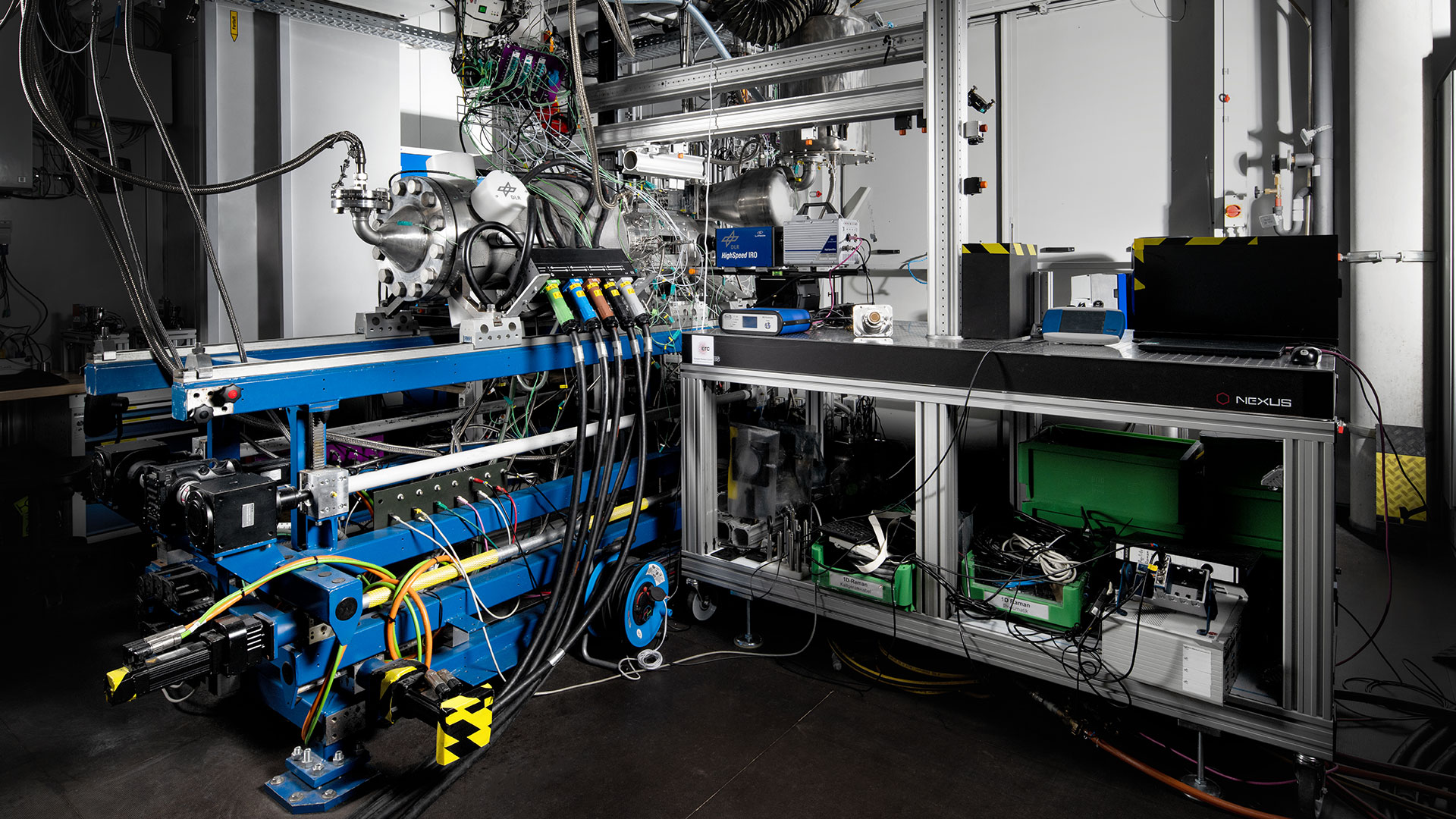 Das Bild zeigt den Hochdrückbrennkammerprüfstand HiPOT. Links ist der blaue, fahrbare Prüfstandsrahmen mit dem darauf befindlichen Luftvorwärmer und der Brennkammer zu sehen. Rechts ein Tisch für die Aufnahme diverser Messtechnik.