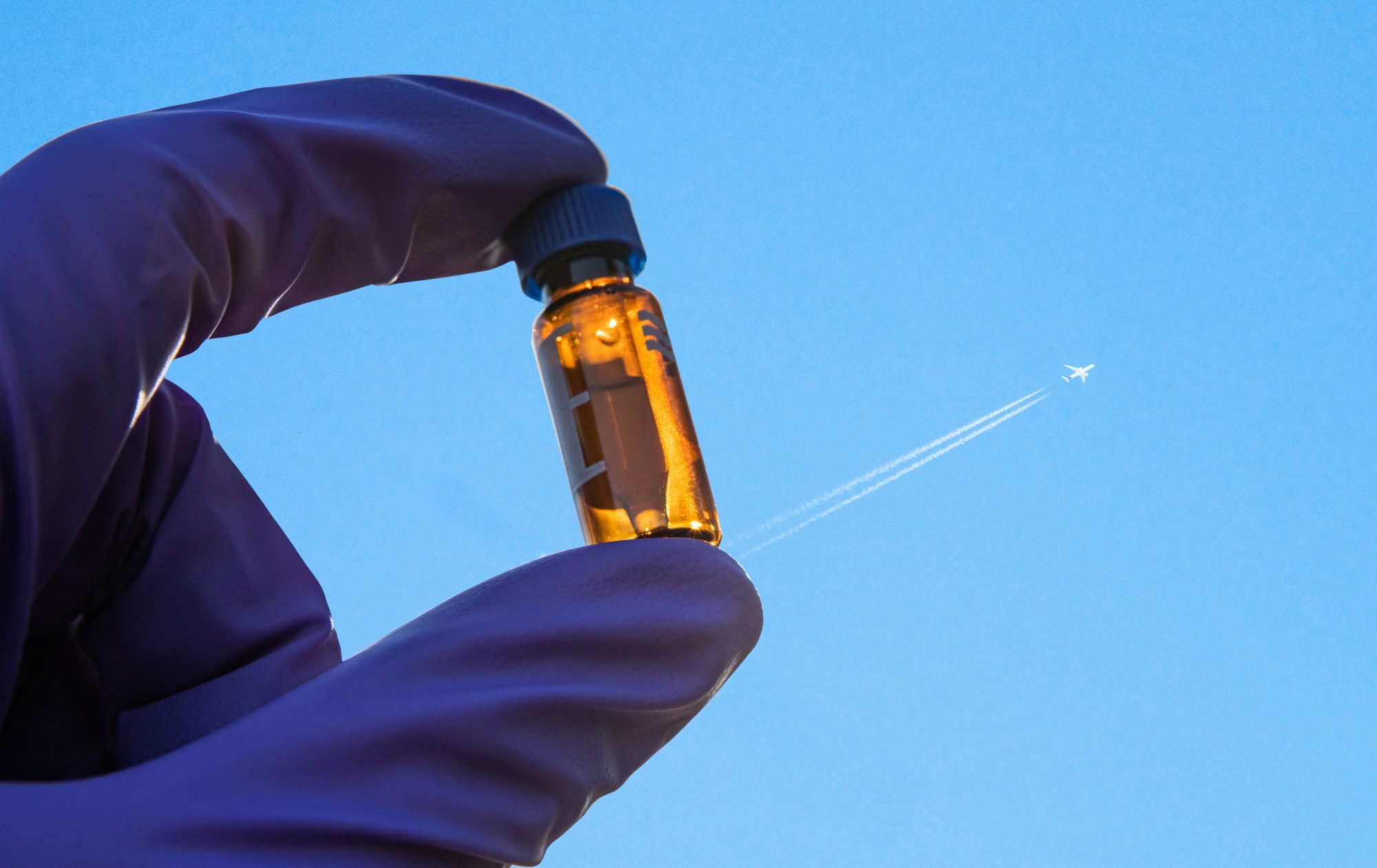 Eine Hand hält ein Probenfläschchen mit Treibstoff gegen den Himmel. Dort ist ein Flugzeug mit Kondensstreifen zu sehen.