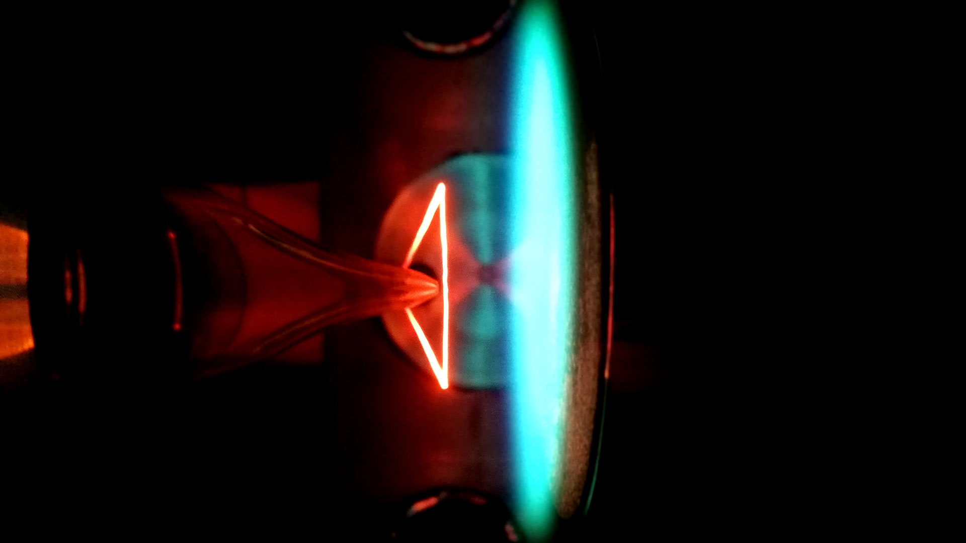 Das Bild zeigt ein reaktionskinetisches Experiment im Strömungsreaktor: das rot glühende Dreieck ist ein Thermoelement, das zur Temperaturbestimmung dient. Das blau glühende Feld daneben eine Flamme im Niederdruckbereich.