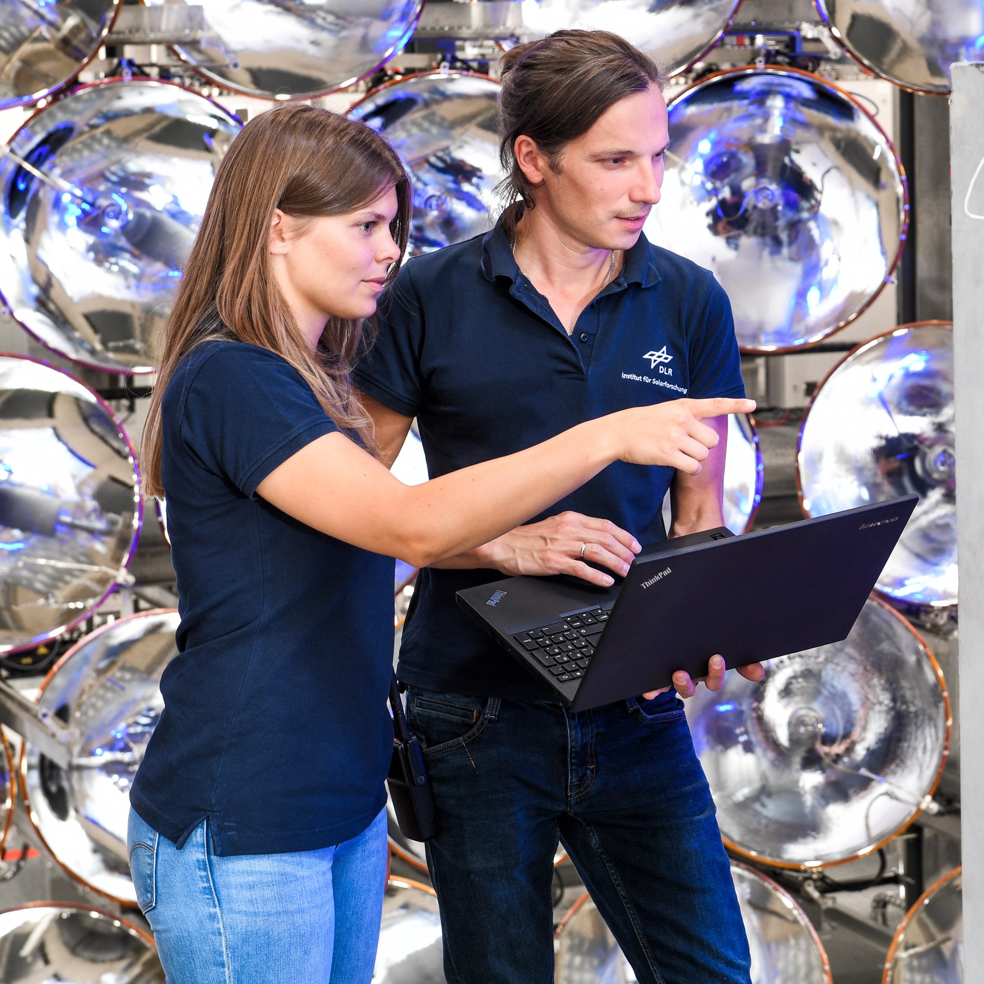 The photograph shows two staff members from the DLR Institute of Solar Research. Both are working on a research project. A woman is standing on the left and a man on the right. The man is resting his laptop on his left forearm and typing with his right hand. The woman is pointing at something with her right hand. High-power radiators can be seen behind them.