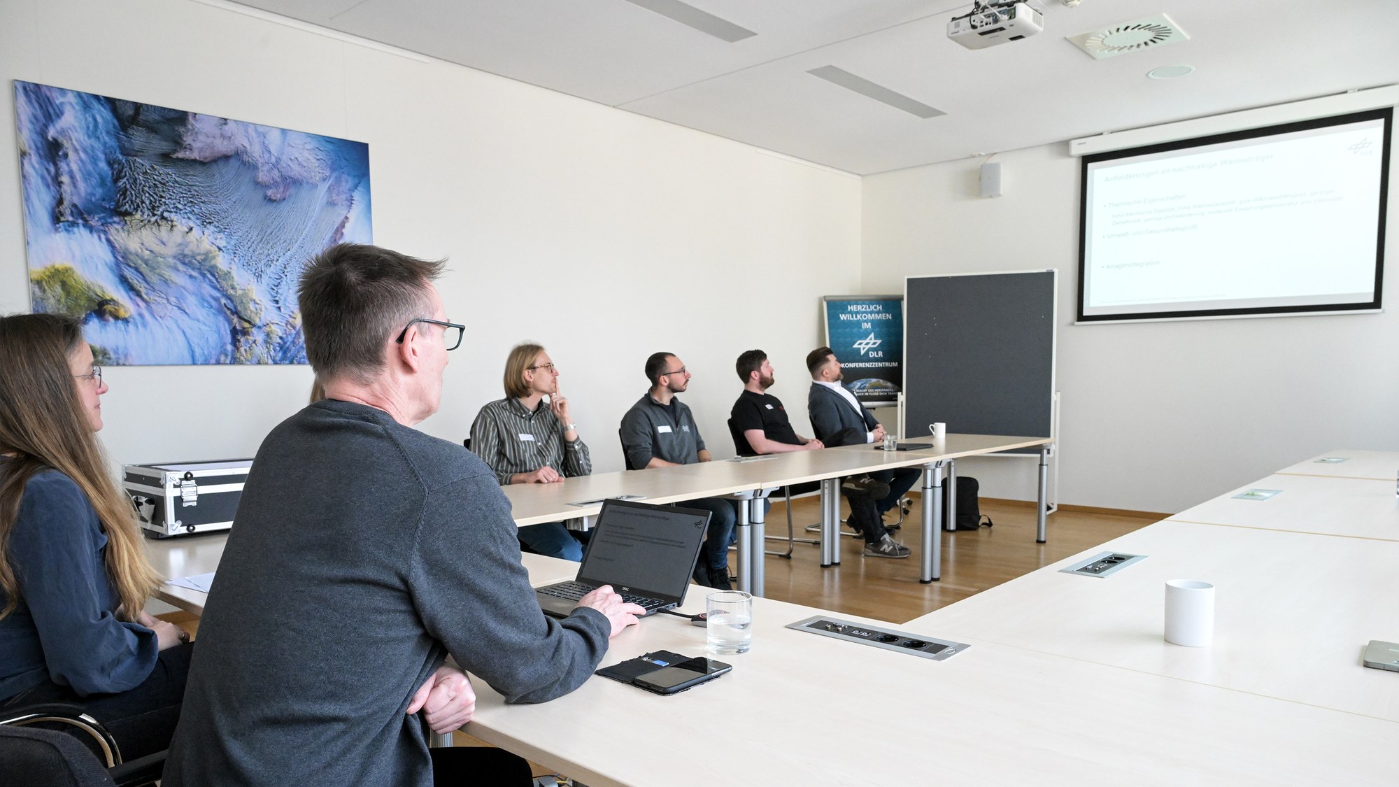 A group is sitting in a seminar room at a U-shaped table and looking at a presentation on a projector.