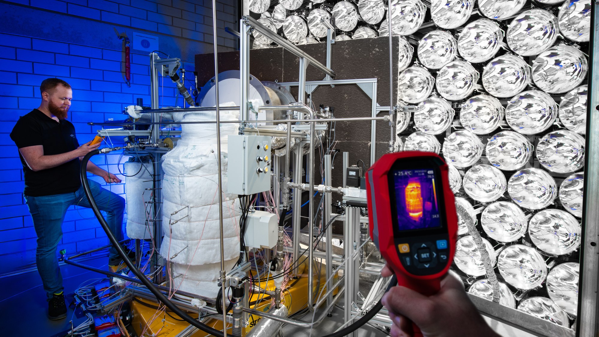 A scientist working on the SOL reactor in Synlight. A hand in the foreground holds a thermal imaging camera on which a heated heat storage tank can be seen in colour.