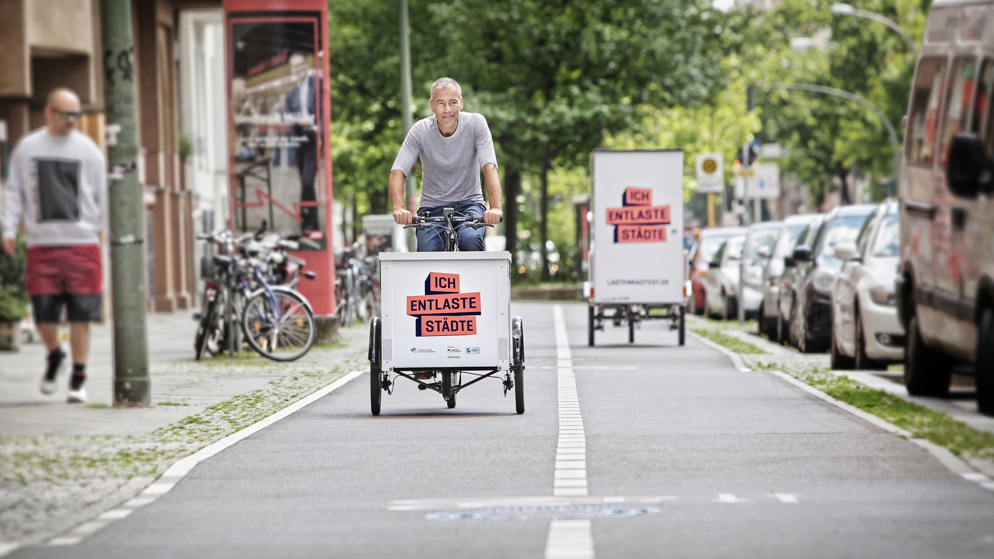 A man rides a cargo bike from the 'Ich entlaste Städte' campaign on a cycle path, with another cargo bike in the background. A passer-by is walking on the pavement next to the cycle path.