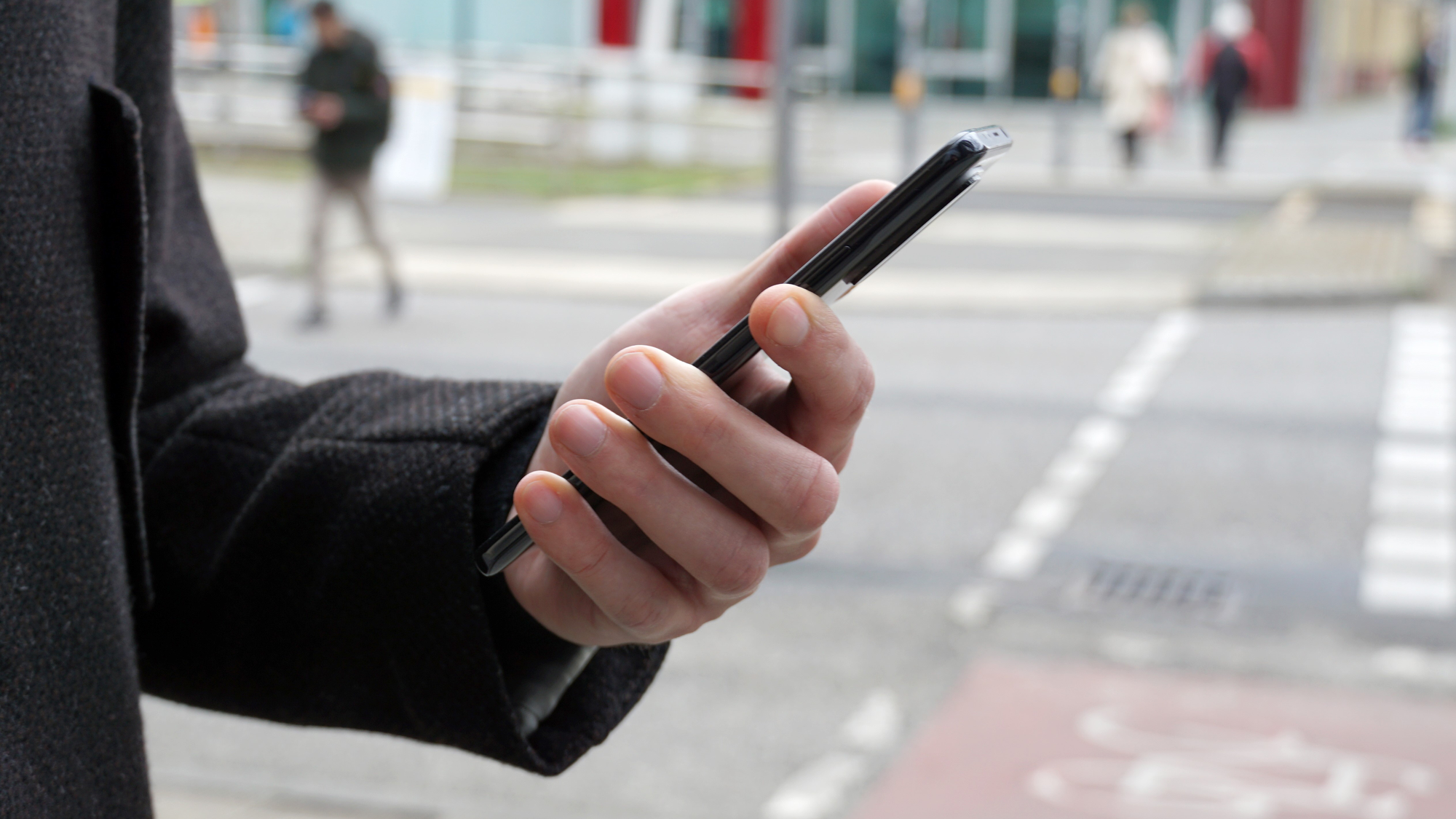 A hand can be seen holding a smartphone. A pedestrian crossing and a cycle path can be seen in the background. Several pedestrians are crossing the road. The content displayed on the smartphone cannot be recognised.