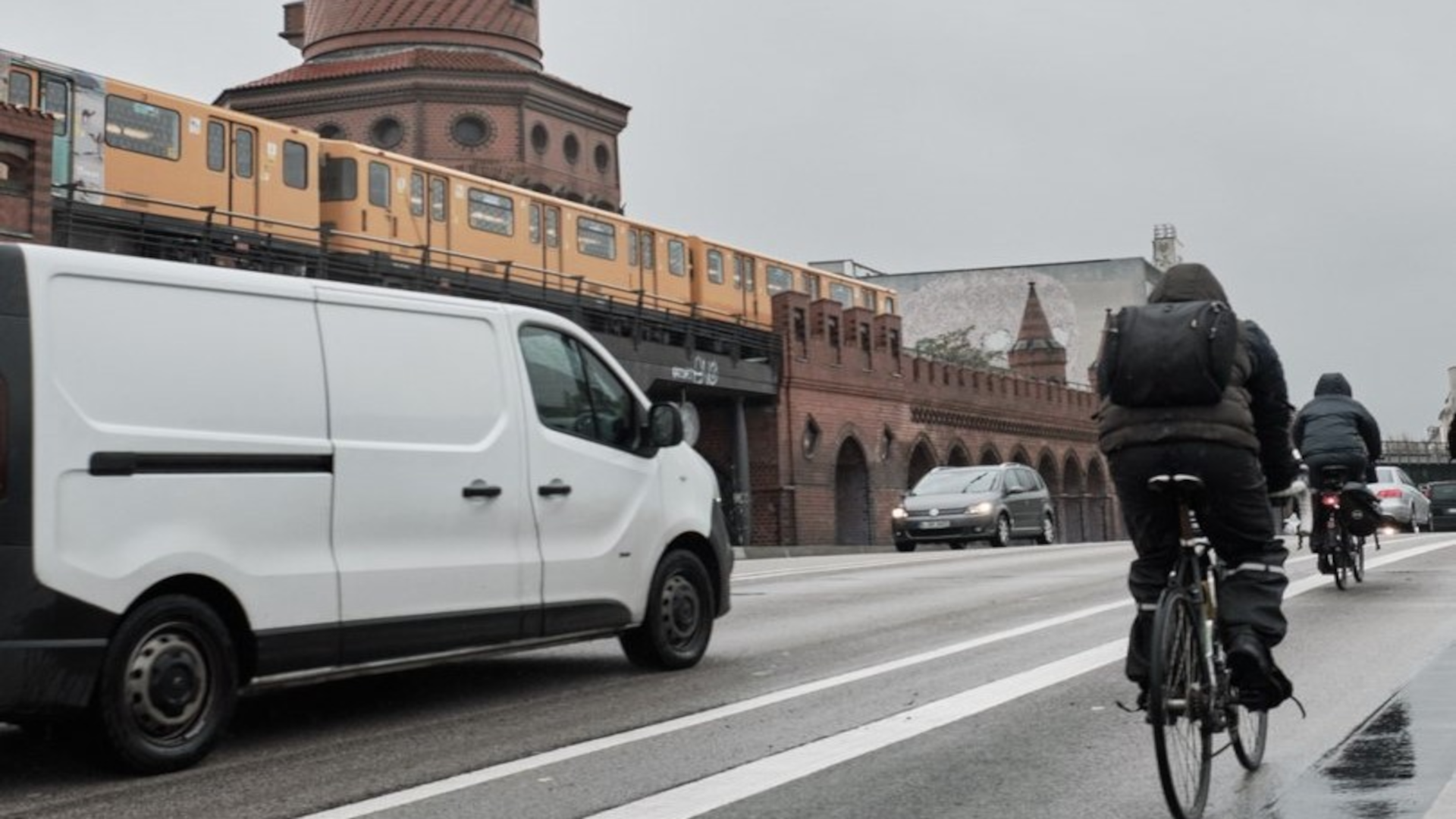 This is a photo of the Oberbaum Bridge in Berlin in rainy weather. An above-ground underground train is travelling across the bridge, as are several cars. Some cyclists in rain gear are riding on the cycle path, which is close to the road.