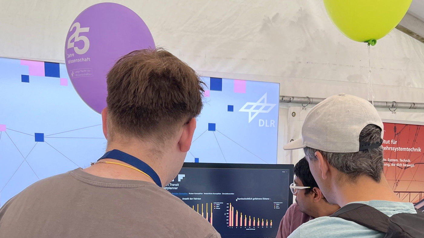 Two men with balloons stand in front of a screen with a scientist.