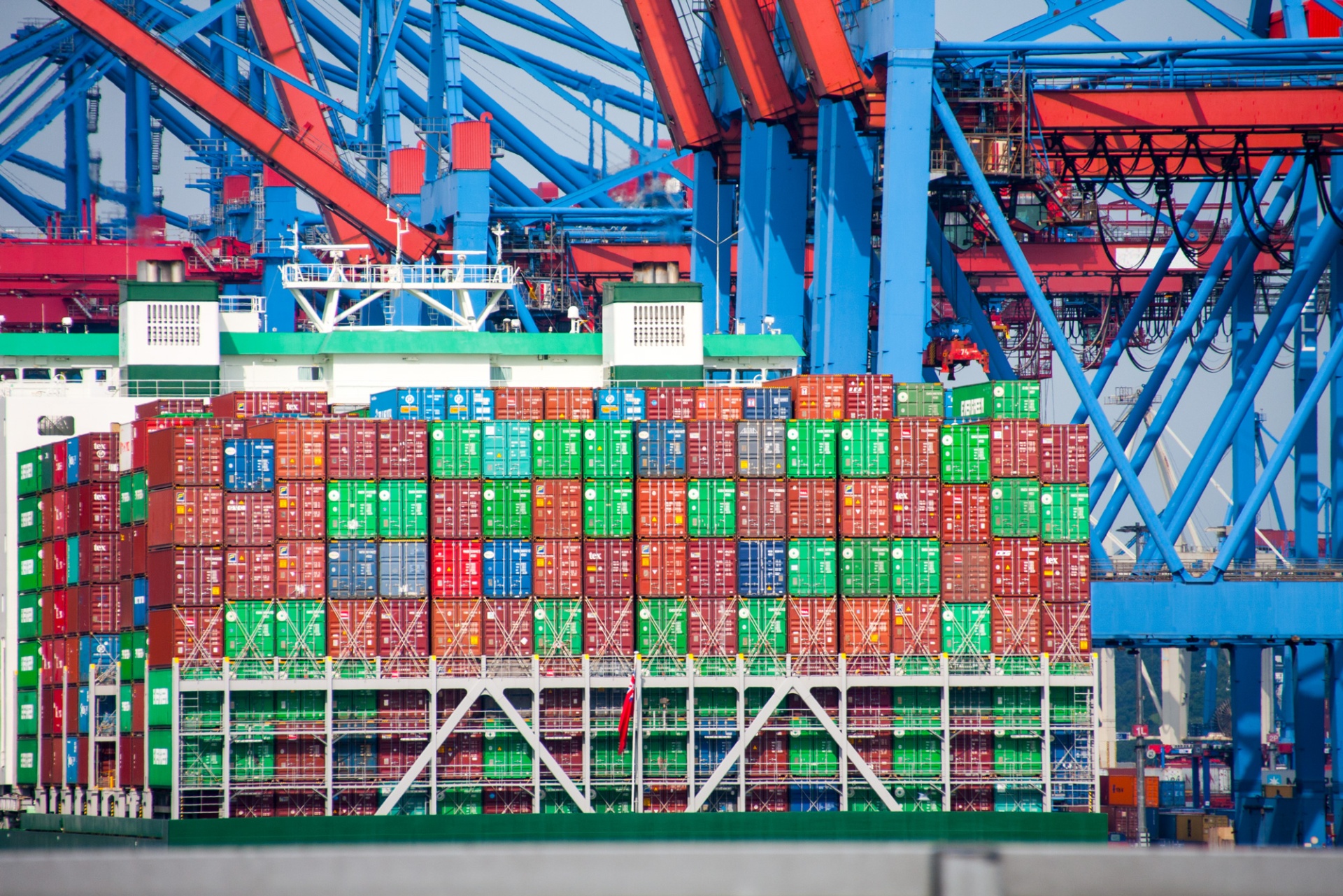 A ship is moored in the harbour basin, loaded with containers. In the background, large cranes are loading even more containers onto the ship.