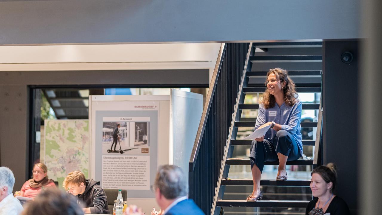 A group of people can be seen sitting and working together in a room for a workshop. The focus is on a lady sitting on a staircase and following the events with interest.
